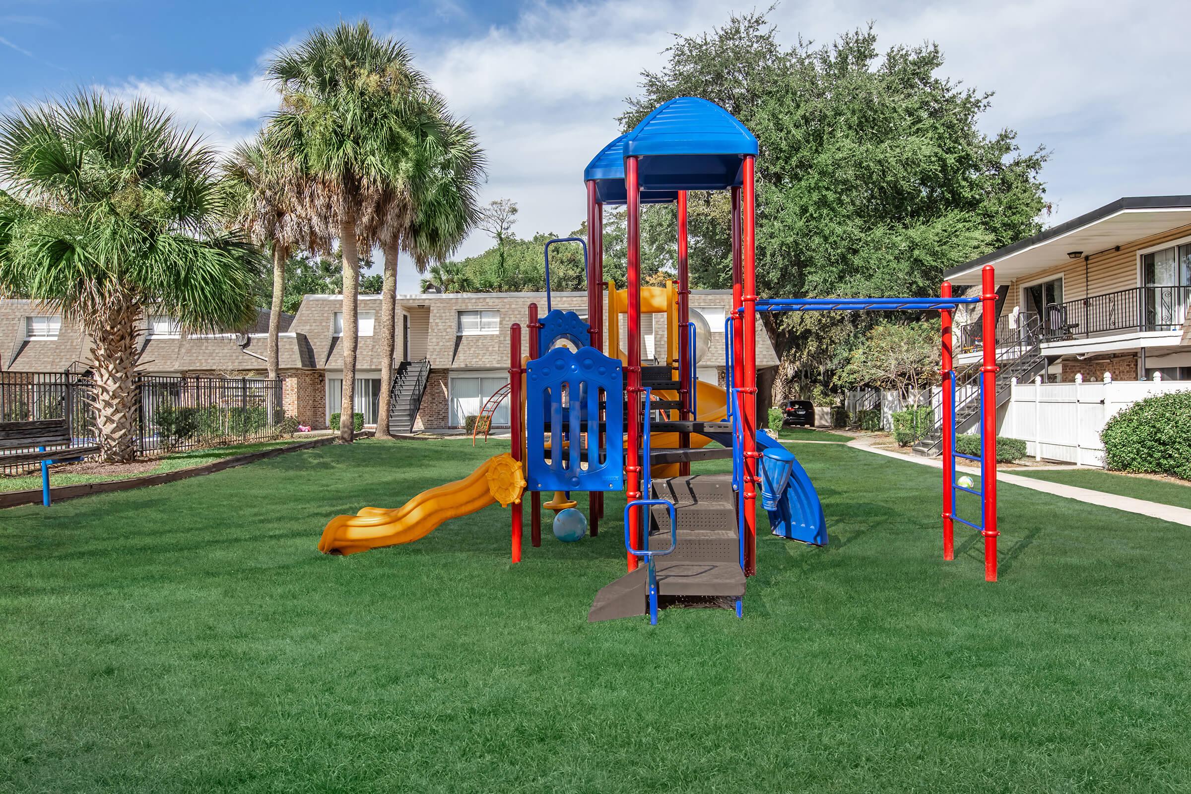 A colorful playground structure with slides, climbing features, and a playhouse, set in a grassy area surrounded by palm trees and residential buildings. The scene is vibrant and inviting, perfect for children's outdoor play.