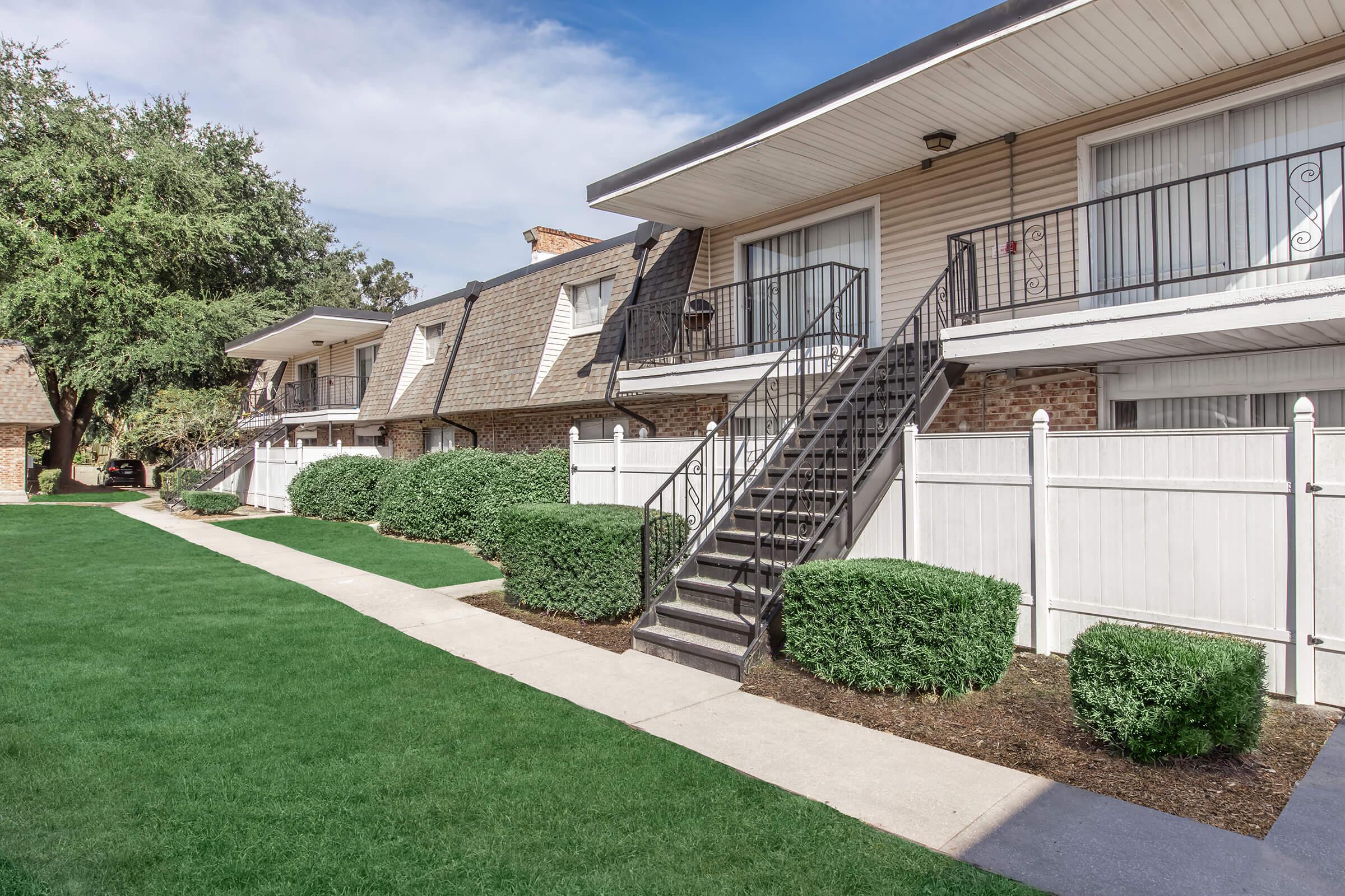 A well-maintained apartment complex with two-story buildings, featuring balconies and a staircase. The green lawn is bordered by neatly trimmed hedges and a white fence. There are trees in the background, and the sky is partly cloudy, creating a pleasant outdoor environment.