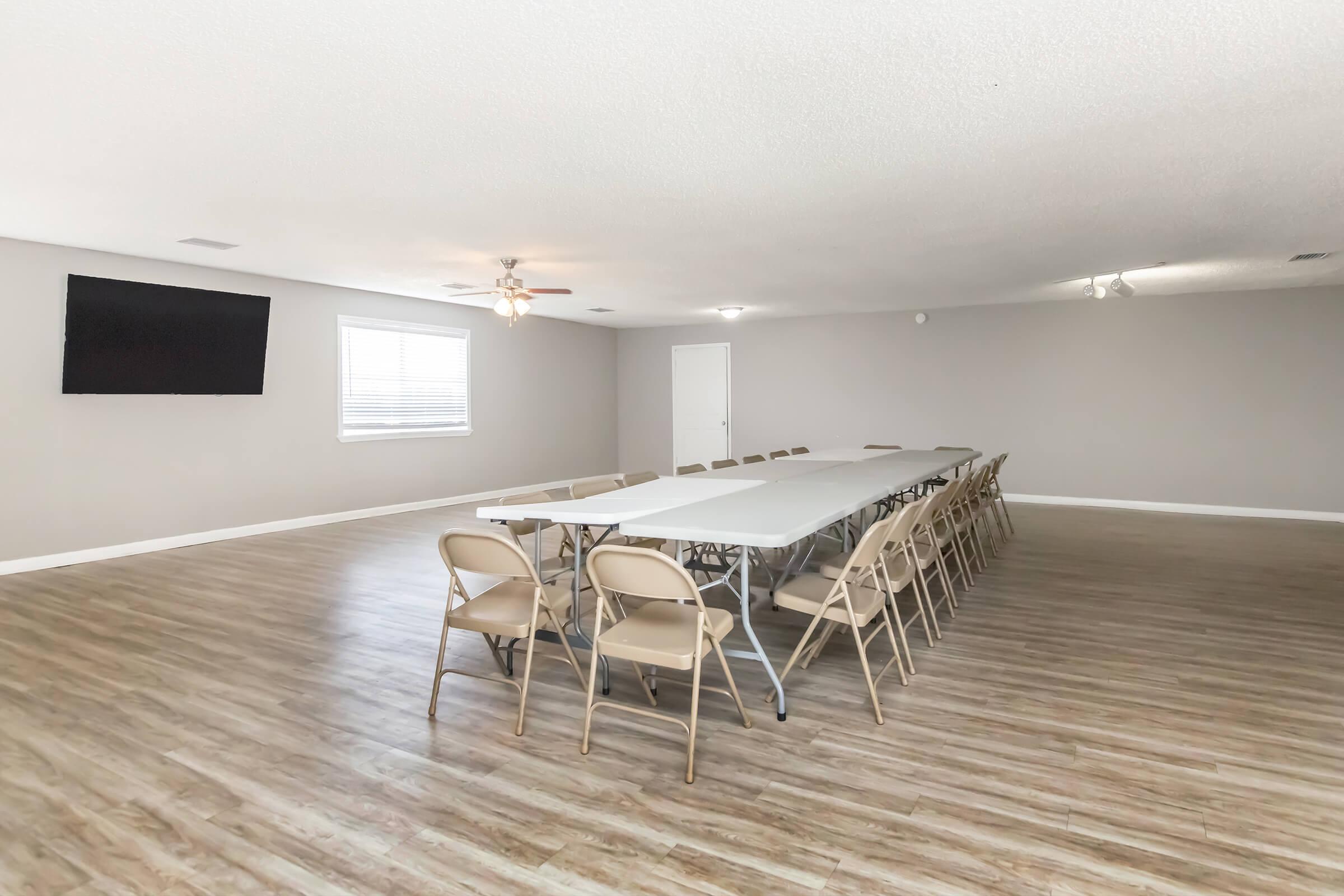A spacious meeting room featuring a long, rectangular table with several folding chairs arranged around it. The room has light wood flooring, light gray walls, and a large flat-screen TV mounted on one wall. Two ceiling fans provide ventilation, and natural light comes from windows with blinds.