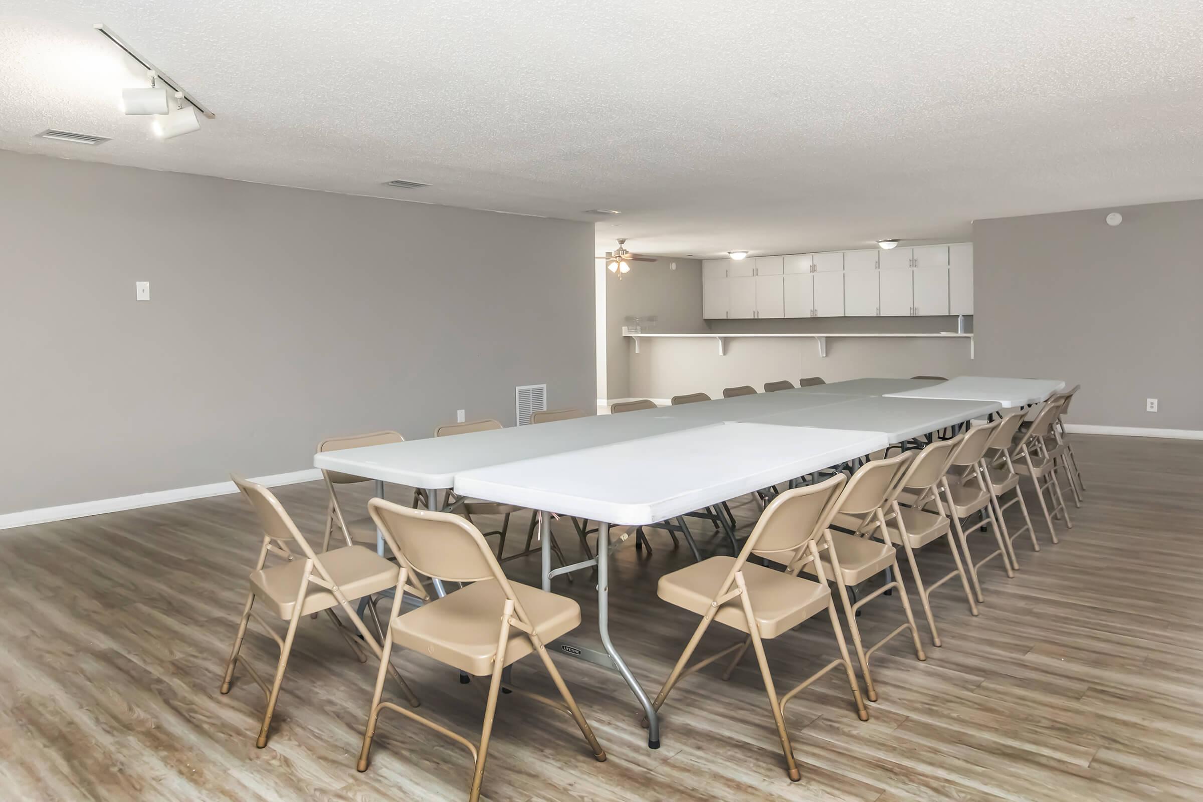 A spacious indoor room featuring multiple folding tables arranged in a U-shape, surrounded by beige chairs. The walls are painted light gray, and there are modern light fixtures on the ceiling. In the background, a kitchenette area is visible with white cabinetry, creating a bright and functional space suitable for meetings or events.