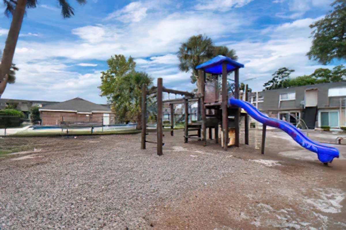 A playground featuring a blue slide, wooden structures, and a sandy ground. Surrounding the playground are trees and residential buildings in the background. The sky is partly cloudy, suggesting a sunny day.