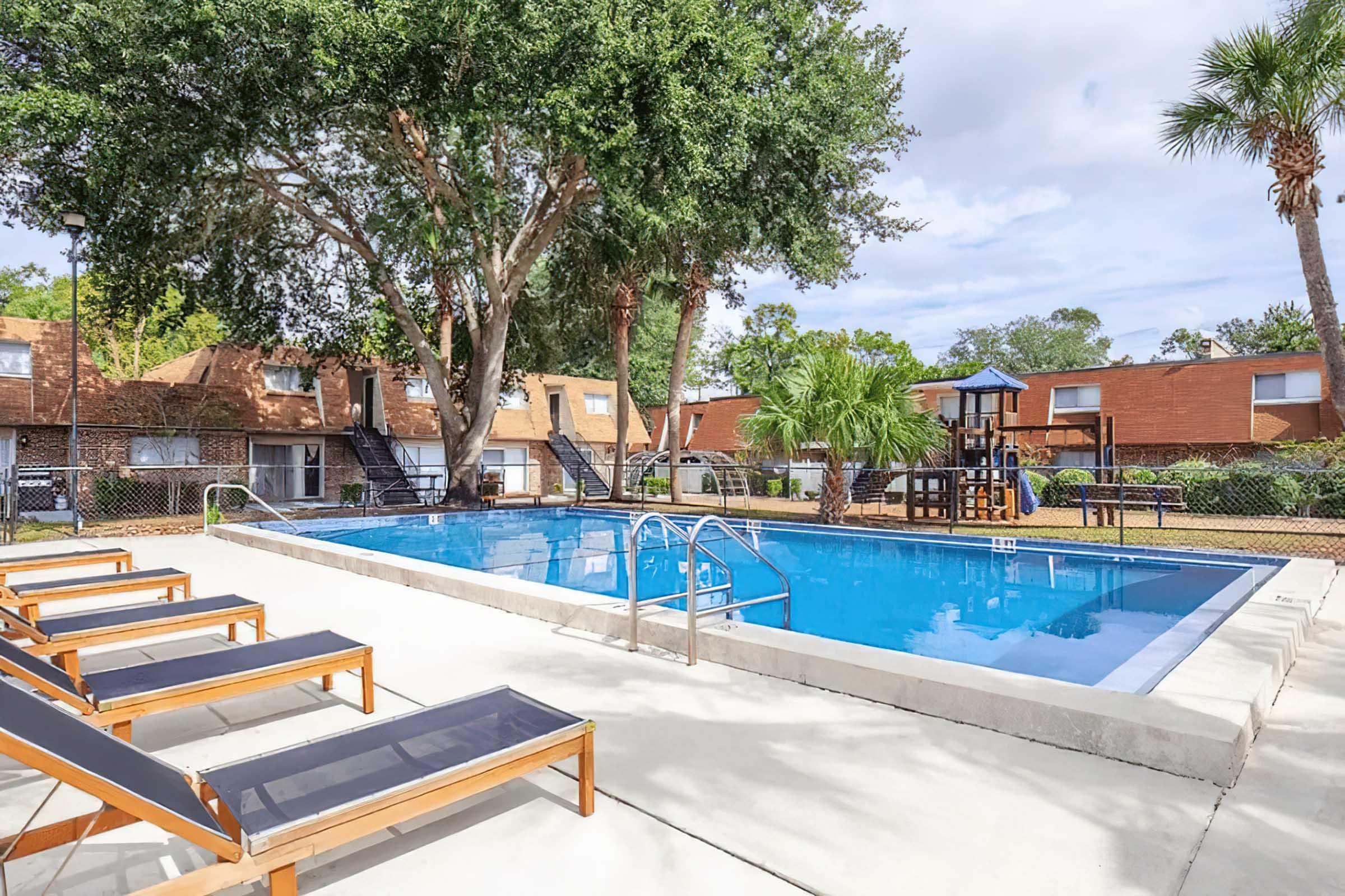 A clear outdoor pool surrounded by lounge chairs, palm trees, and colorful townhouses in the background. The area is well-maintained with a playground visible nearby, creating a relaxing and inviting atmosphere.