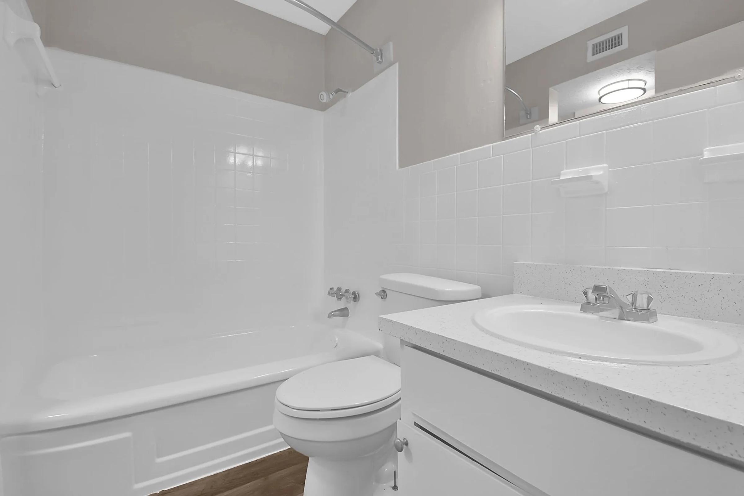 A clean and modern bathroom featuring a white bathtub, toilet, and a sink with a granite countertop. Light tiles are used for the shower area, and a large mirror is above the sink. Natural light enters through the window, giving the space an airy feel.