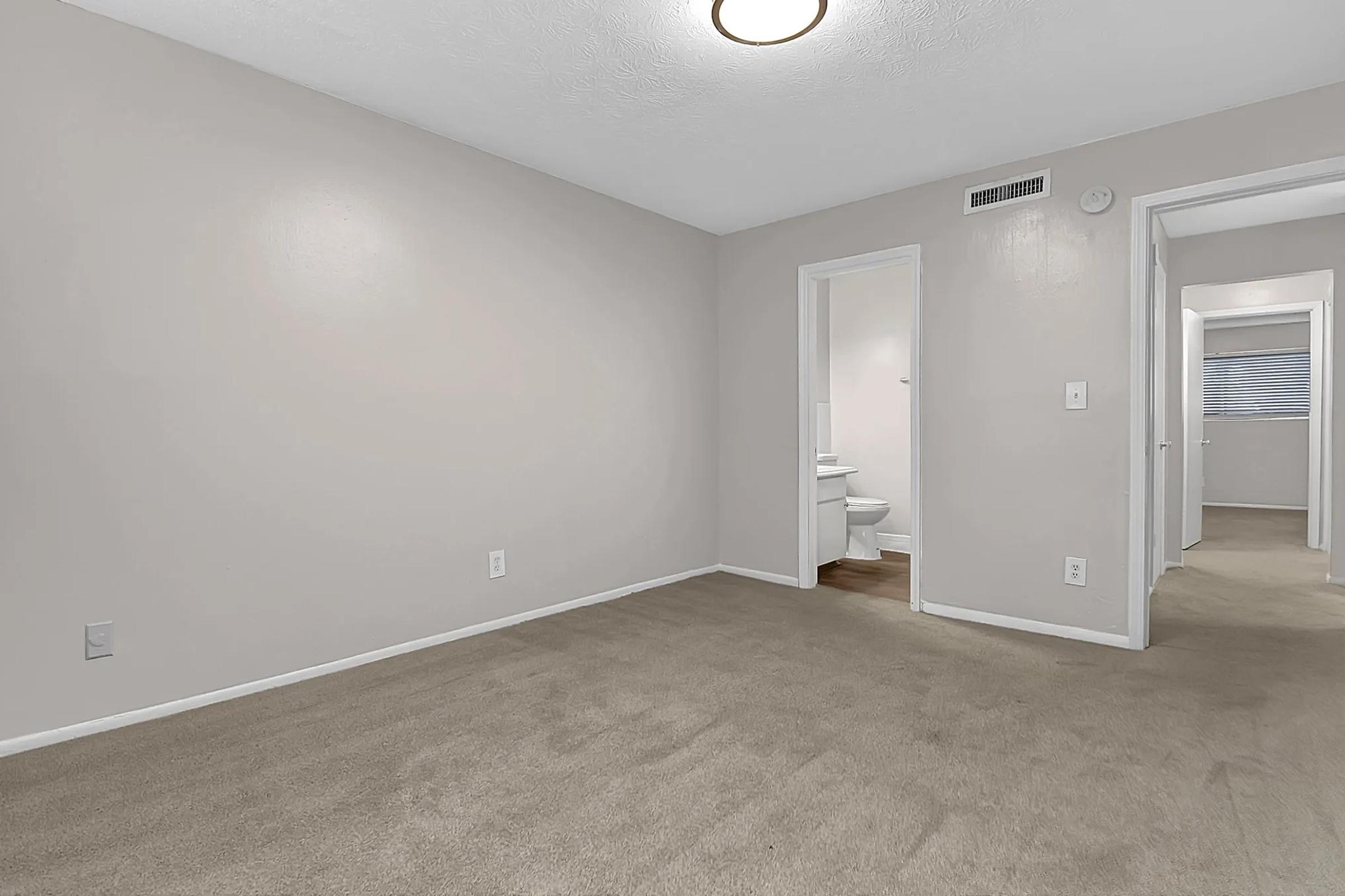 A spacious, empty room with light beige walls and carpet. There are two doorways leading to other areas, one of which appears to lead to a bathroom. The ceiling has a simple light fixture, and there is a window on the far wall, allowing natural light to enter.