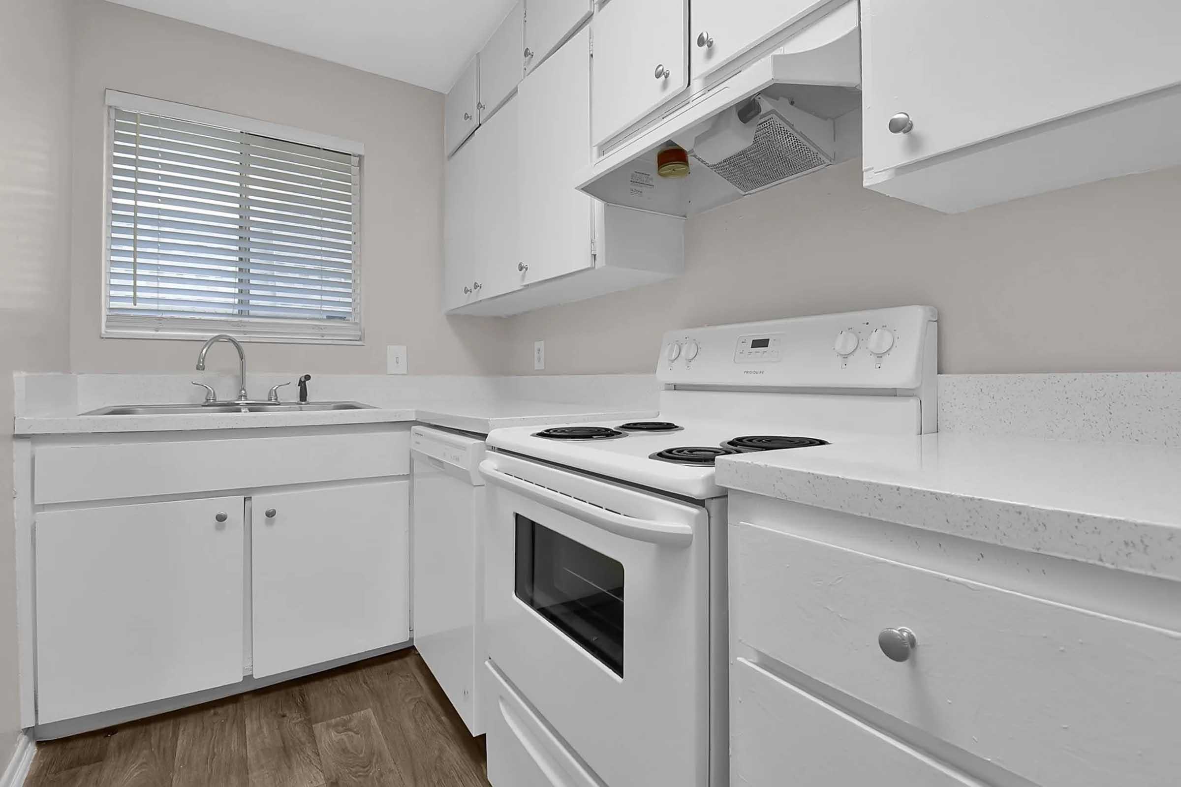 A bright, modern kitchen featuring white cabinets, a double sink under a window with horizontal blinds, an electric stove, and an oven. The countertops are light-colored, complementing the overall clean and spacious design of the kitchen.