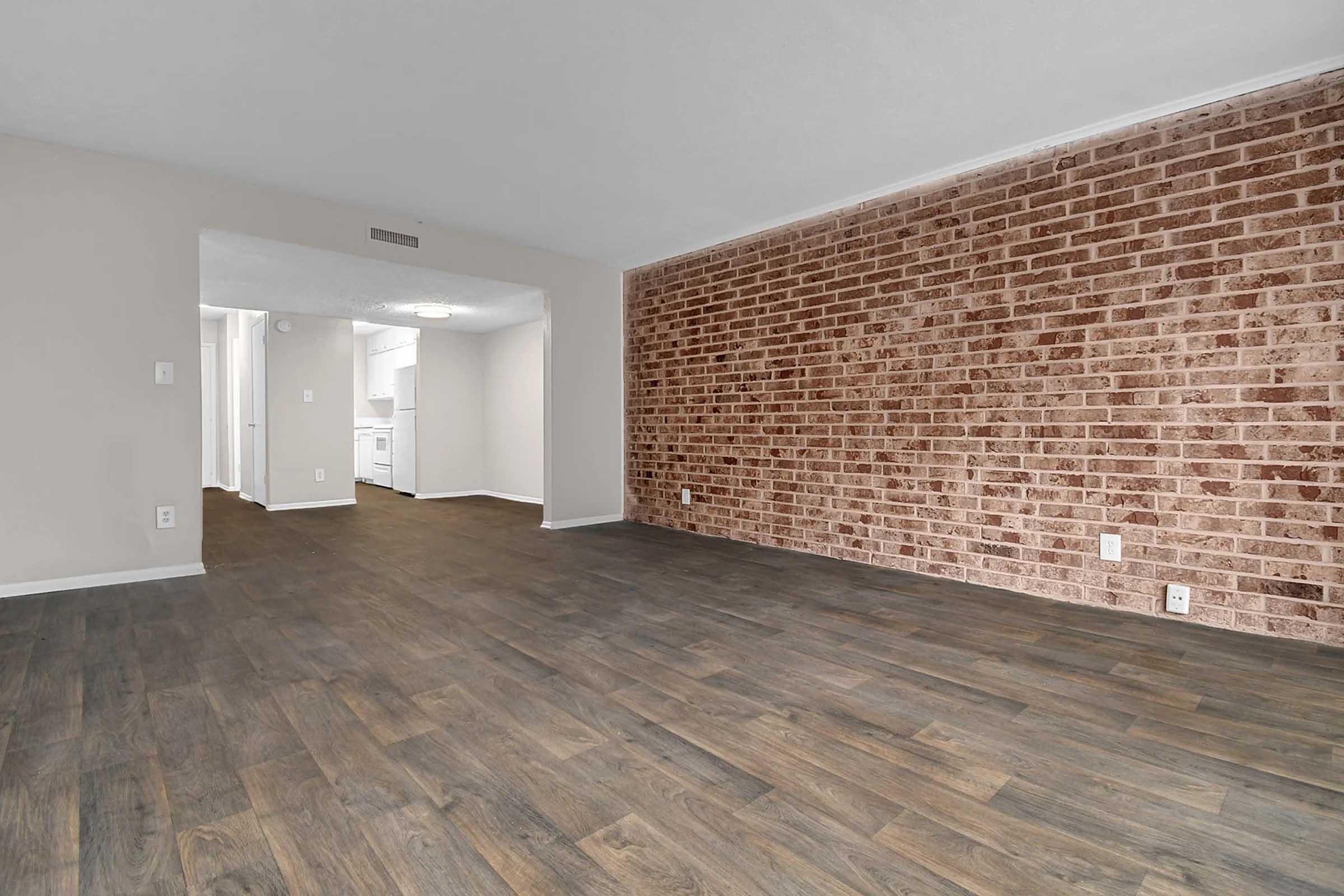 A spacious living area featuring a wood-like floor, a brick accent wall on one side, and an opening leading to a kitchen area. The walls are painted in a light color, contributing to a bright and airy atmosphere.