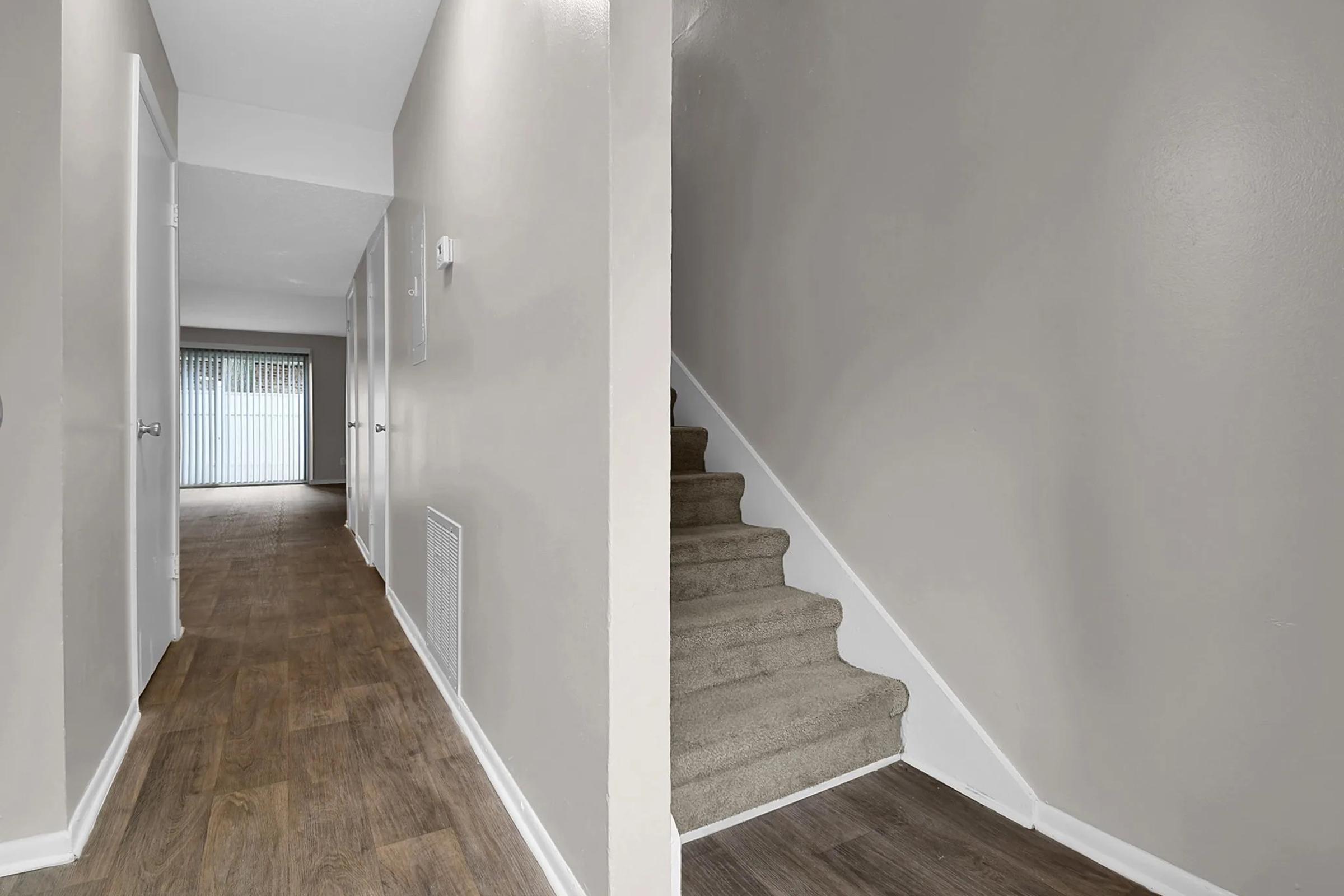 A clean and modern interior space featuring a hallway with two closed doors on the left and a staircase on the right. The walls are painted a light gray, and the flooring has a wood-like appearance. Natural light filters in from a window at the end of the hallway.