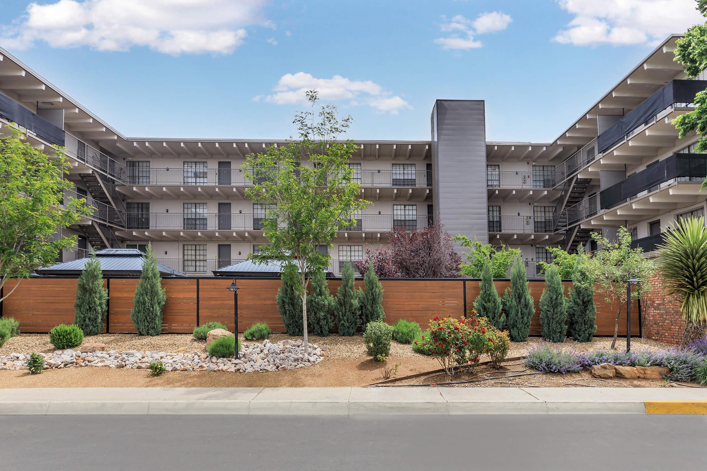 A modern apartment complex with multiple stories, surrounded by well-maintained landscaping. The outdoor area features trees, bushes, and flowers, along with decorative rocks and a wooden fence. The building has balconies and large windows, set against a clear blue sky.
