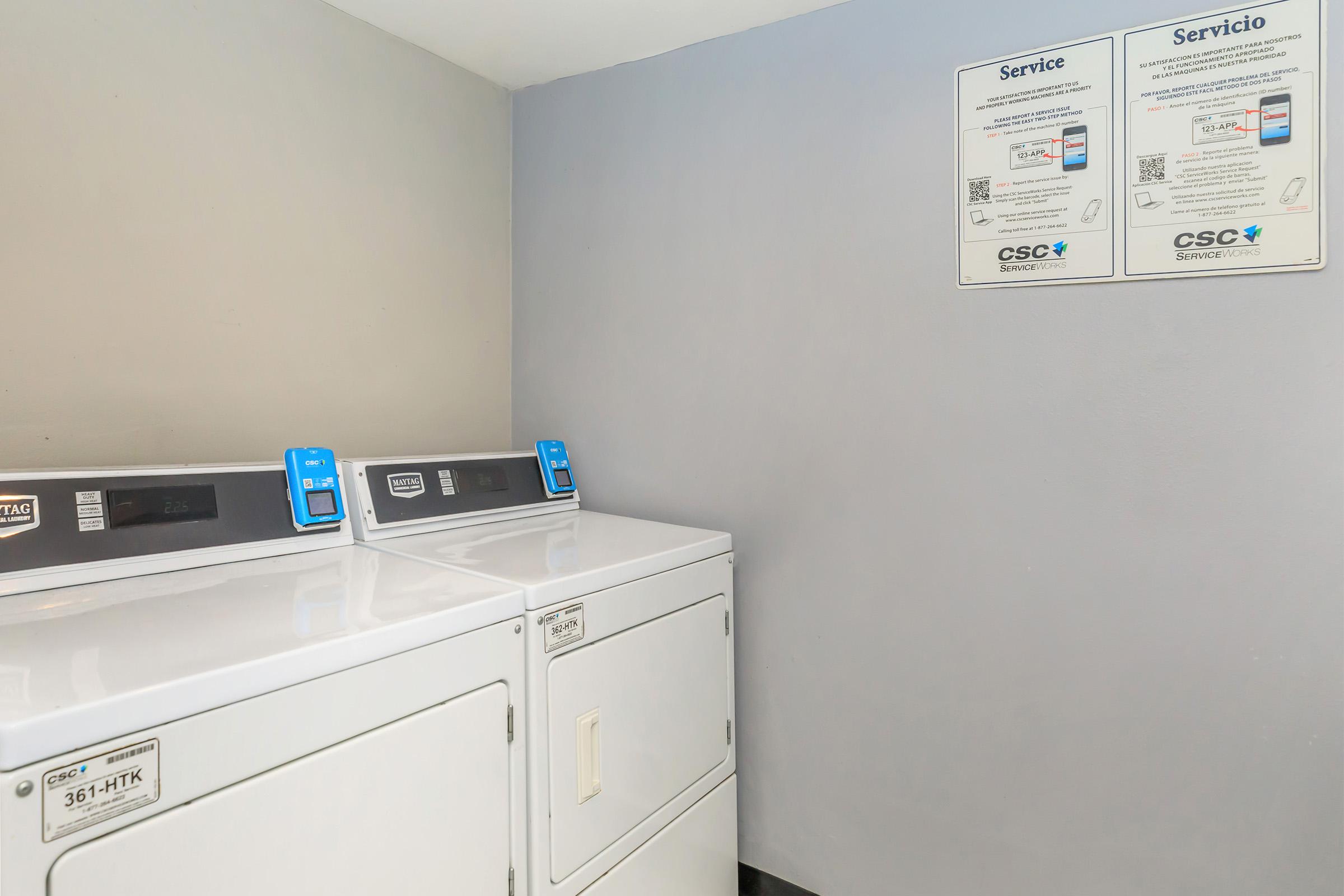 A clean laundry room featuring two stacked washing machines with digital controls. On the wall, there are bilingual service information signs in English and Spanish, displaying QR codes for additional assistance. The background is a simple gray wall, creating a modern and tidy atmosphere.