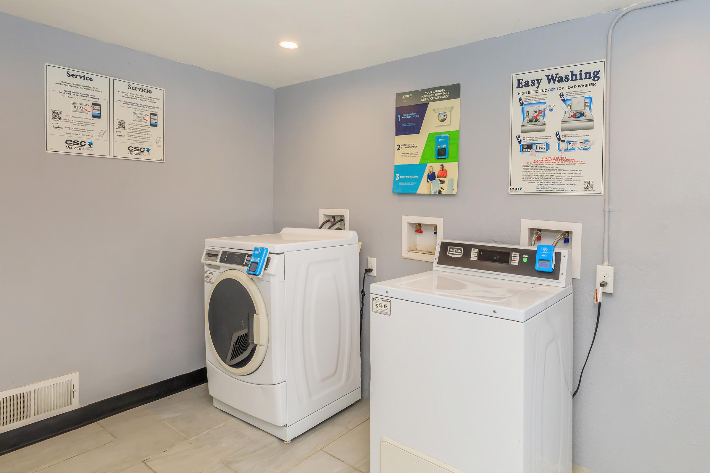 A laundry room featuring a white front-loading dryer and a top-loading washing machine, both equipped with digital controls. The walls are painted light gray, and there are informational signs about laundry services and instructions nearby. The flooring is tiled.