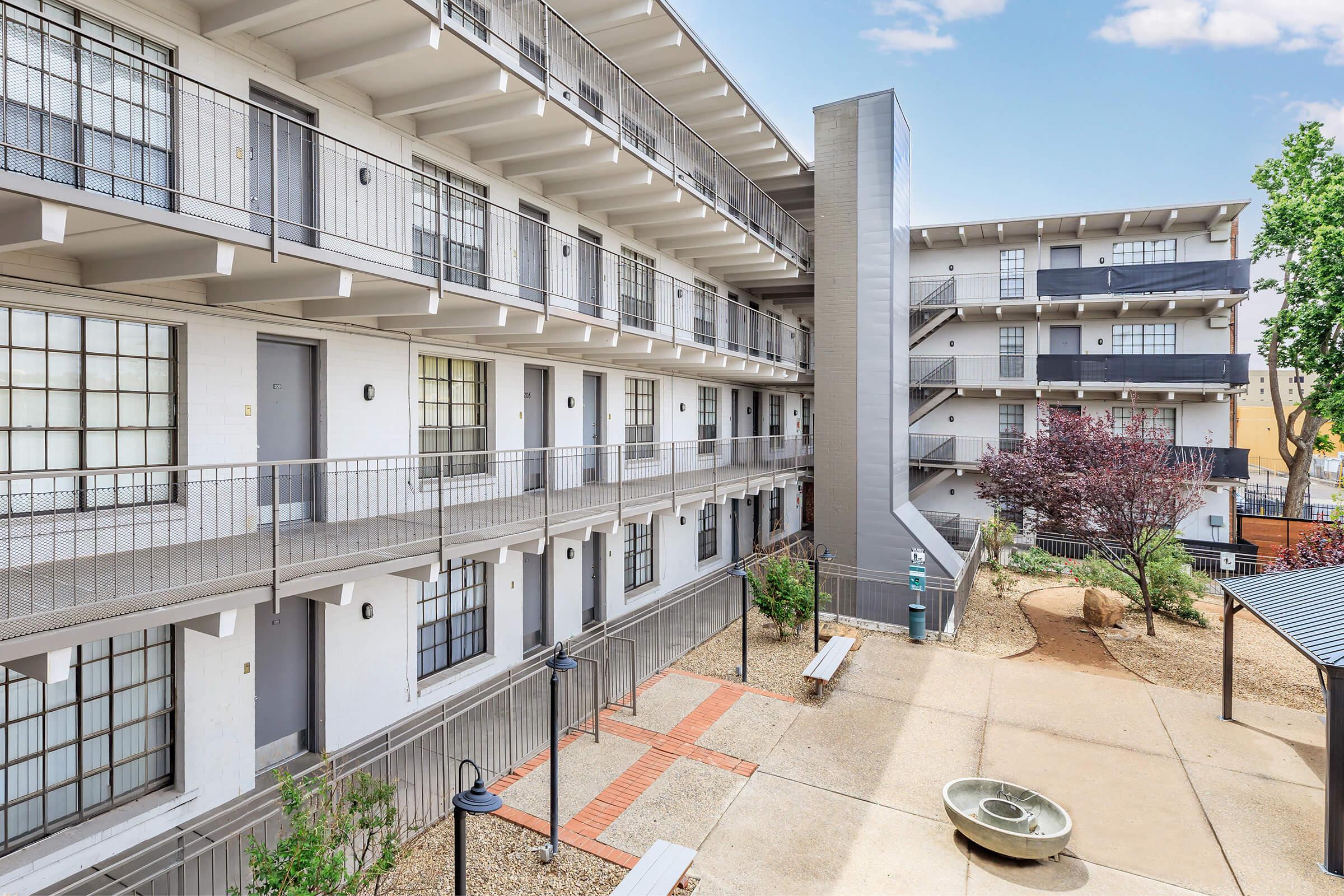 A view of a multi-story building with a modern architectural design, featuring multiple balconies and rows of windows. The courtyard includes benches, landscaping, and a decorative water feature, providing a communal outdoor space for residents.