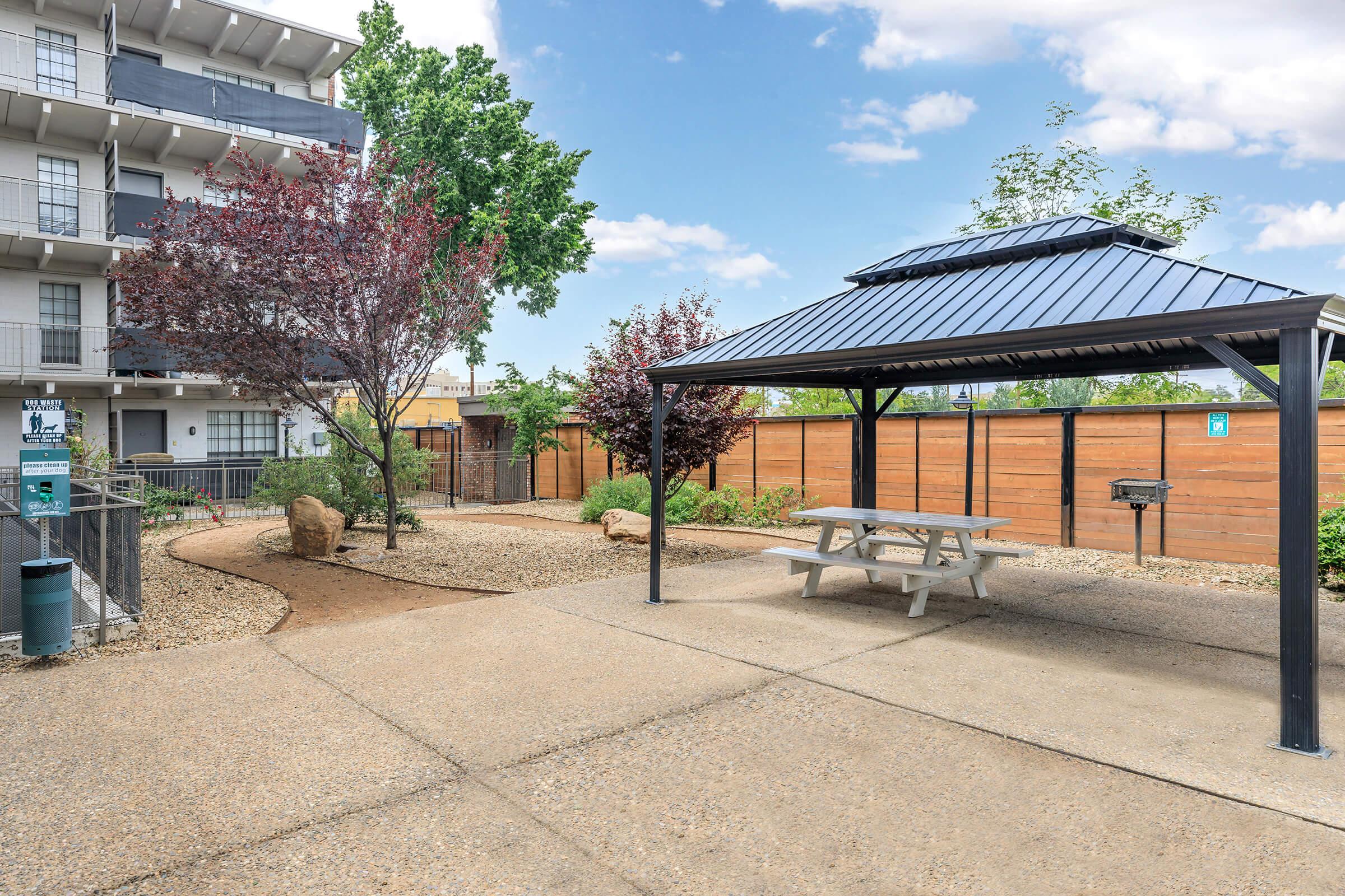 A landscaped outdoor area featuring a picnic table under a metal gazebo, surrounded by small trees and rocks. The space includes a barbecue grill and a pathway, with a modern multi-story building visible in the background. The setting appears inviting and well-maintained.