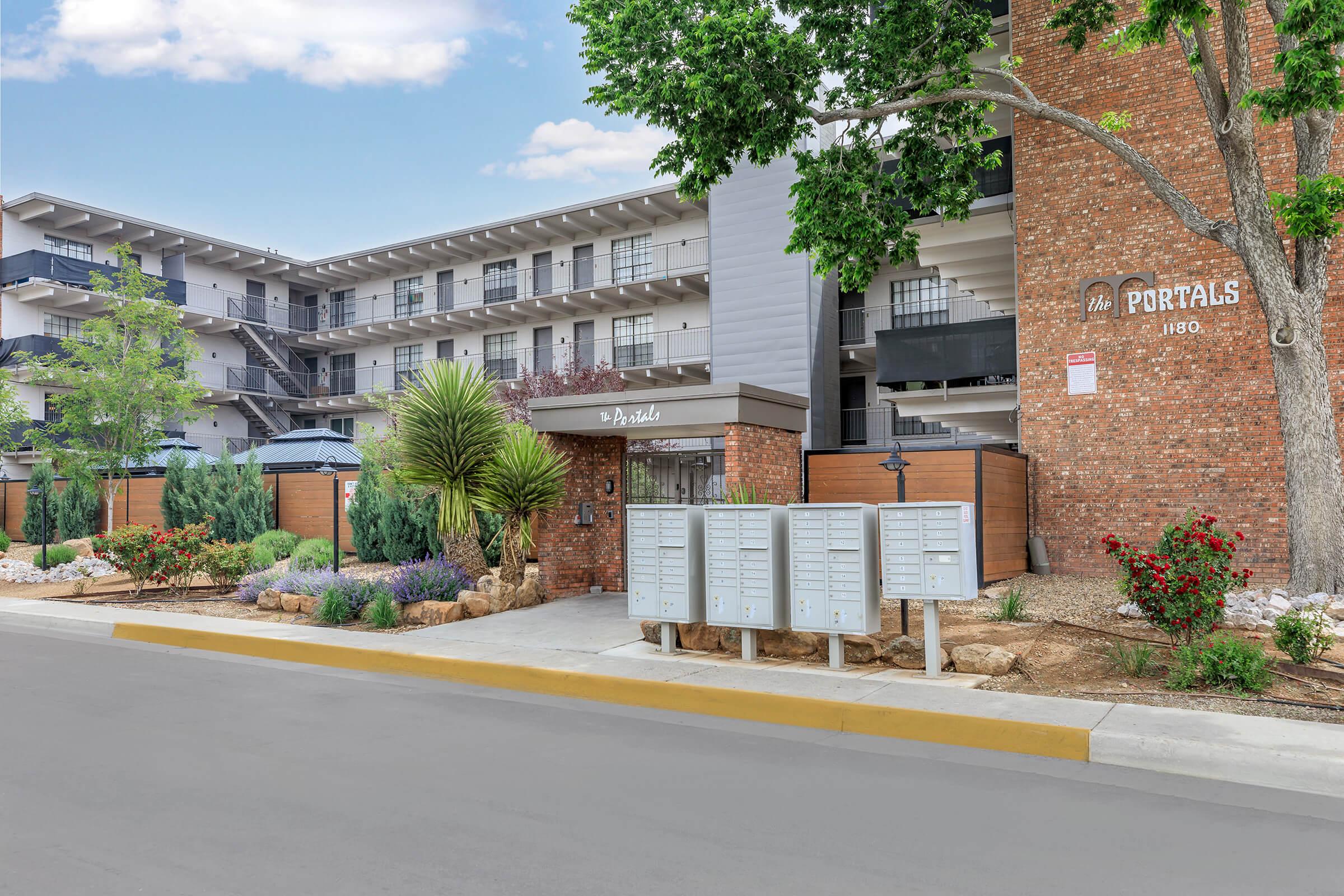 Exterior view of a modern apartment building named "The Portals" at 1180, featuring a brick facade and landscaped surroundings. Mailboxes are situated at the front, with trees and shrubs enhancing the entrance area. The building has multiple stories and balconies visible. The sky is clear and blue.