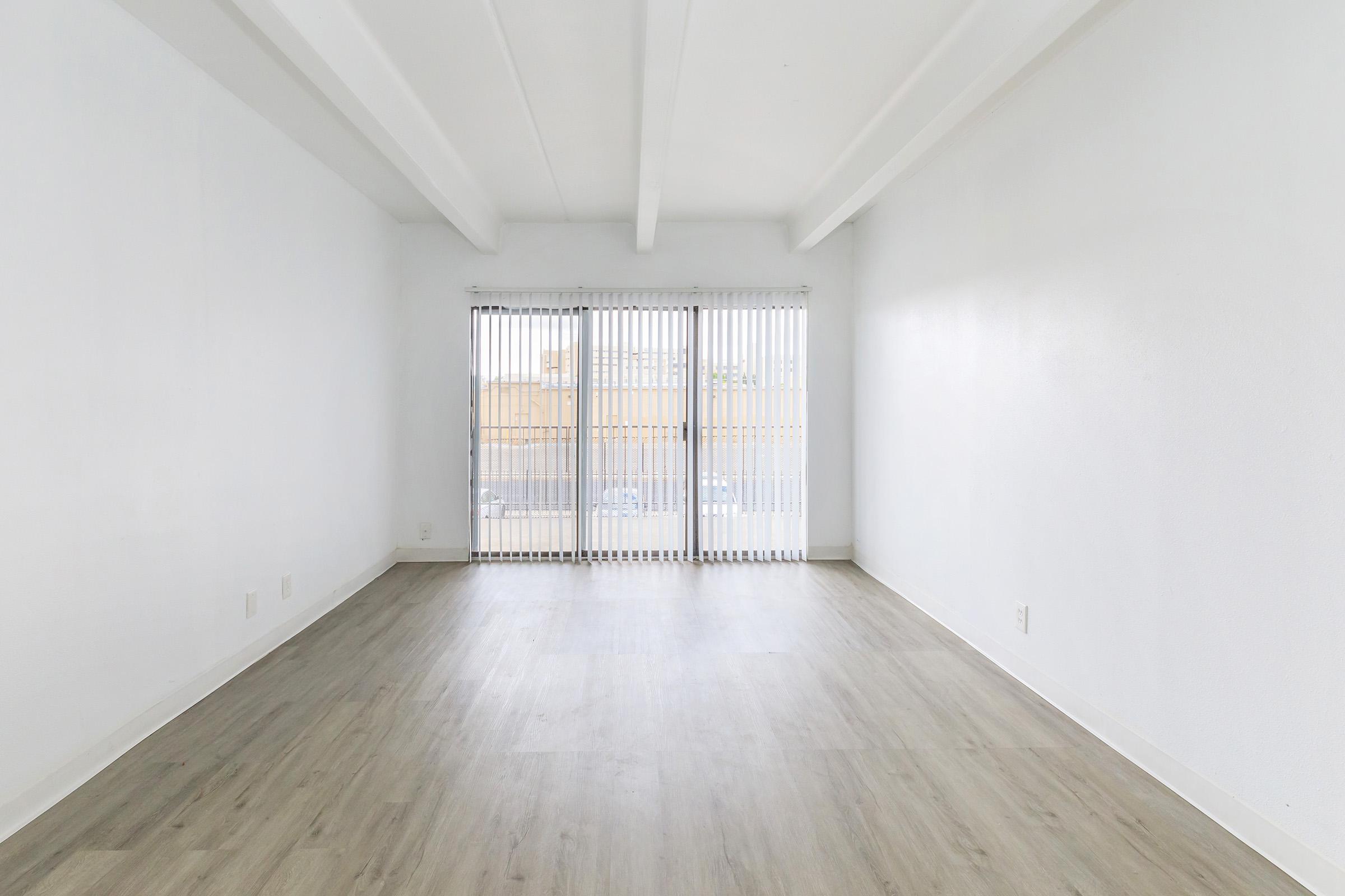 A spacious, minimally furnished room with white walls and wooden flooring. Large sliding glass doors with vertical blinds open to a bright exterior view. The area is well-lit, creating an airy and open atmosphere.