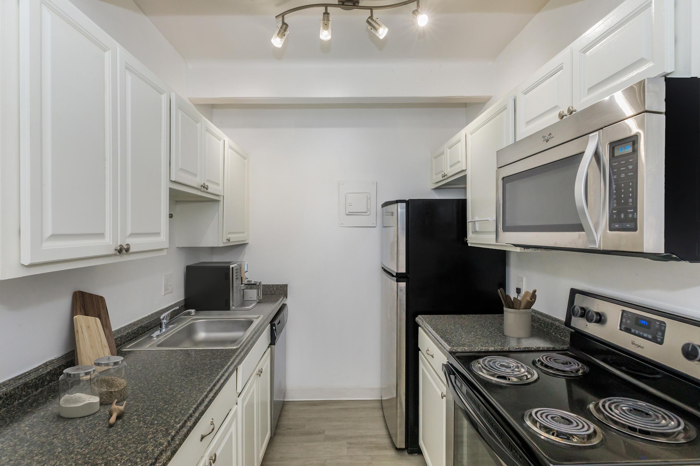 Modern kitchen featuring white cabinets, stainless steel appliances, and a dark granite countertop. The space includes a sink, microwave, and an electric stove with four burners. There are also cooking utensils and a cutting board on the counter, with ceiling lights providing bright illumination.