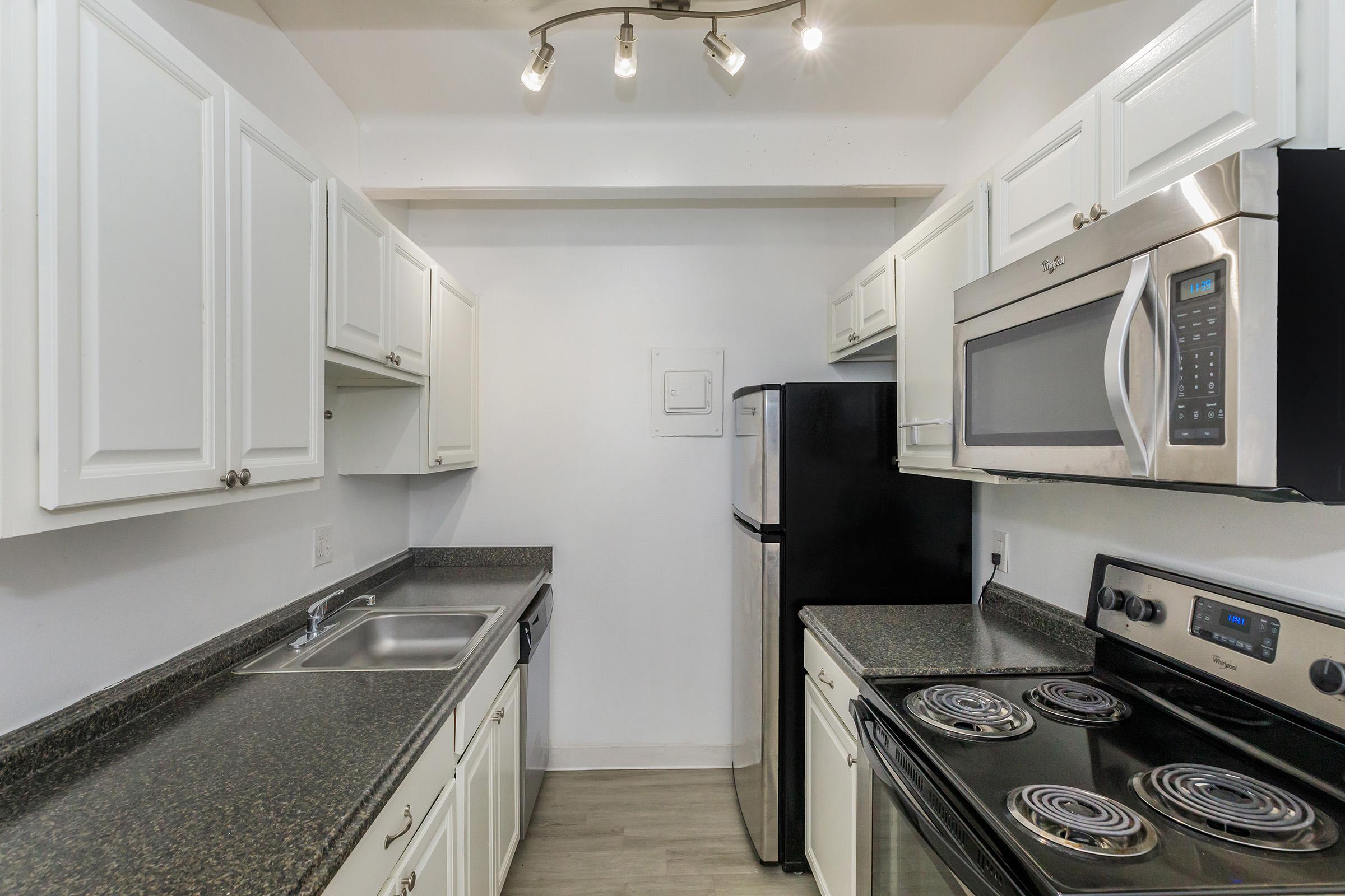 A modern kitchen featuring white cabinetry, a stainless steel refrigerator, a built-in microwave, and a black stove with an oven. The countertop is a dark granite, and there are under-cabinet lights illuminating the space. The kitchen is compact and organized, with a sink installed.