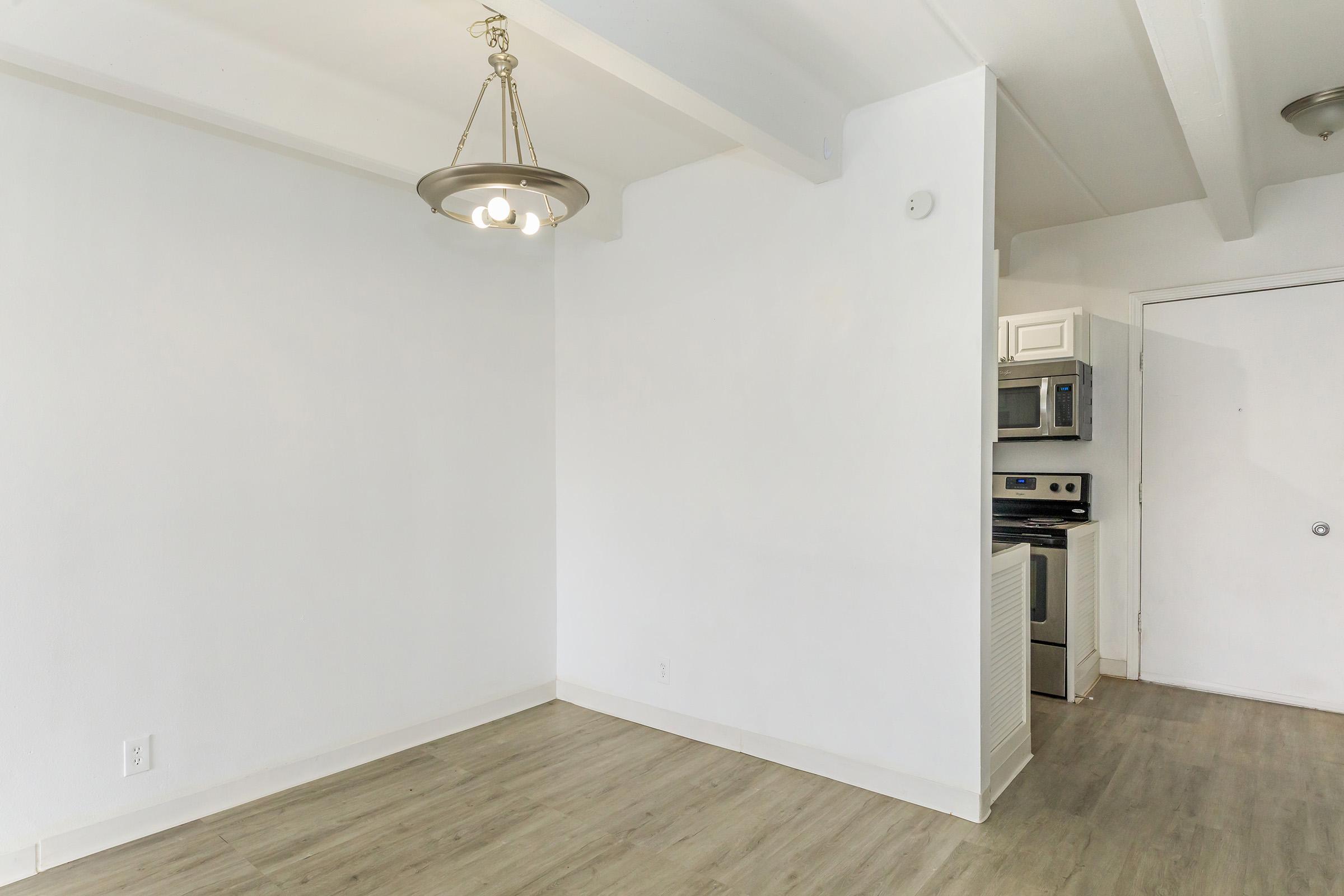Empty interior space featuring light-colored walls and a wooden floor. A simple pendant light hangs from the ceiling. On the right side, there's a door and an adjacent kitchen area with modern appliances including a stove and microwave visible. The space is well-lit and appears inviting.