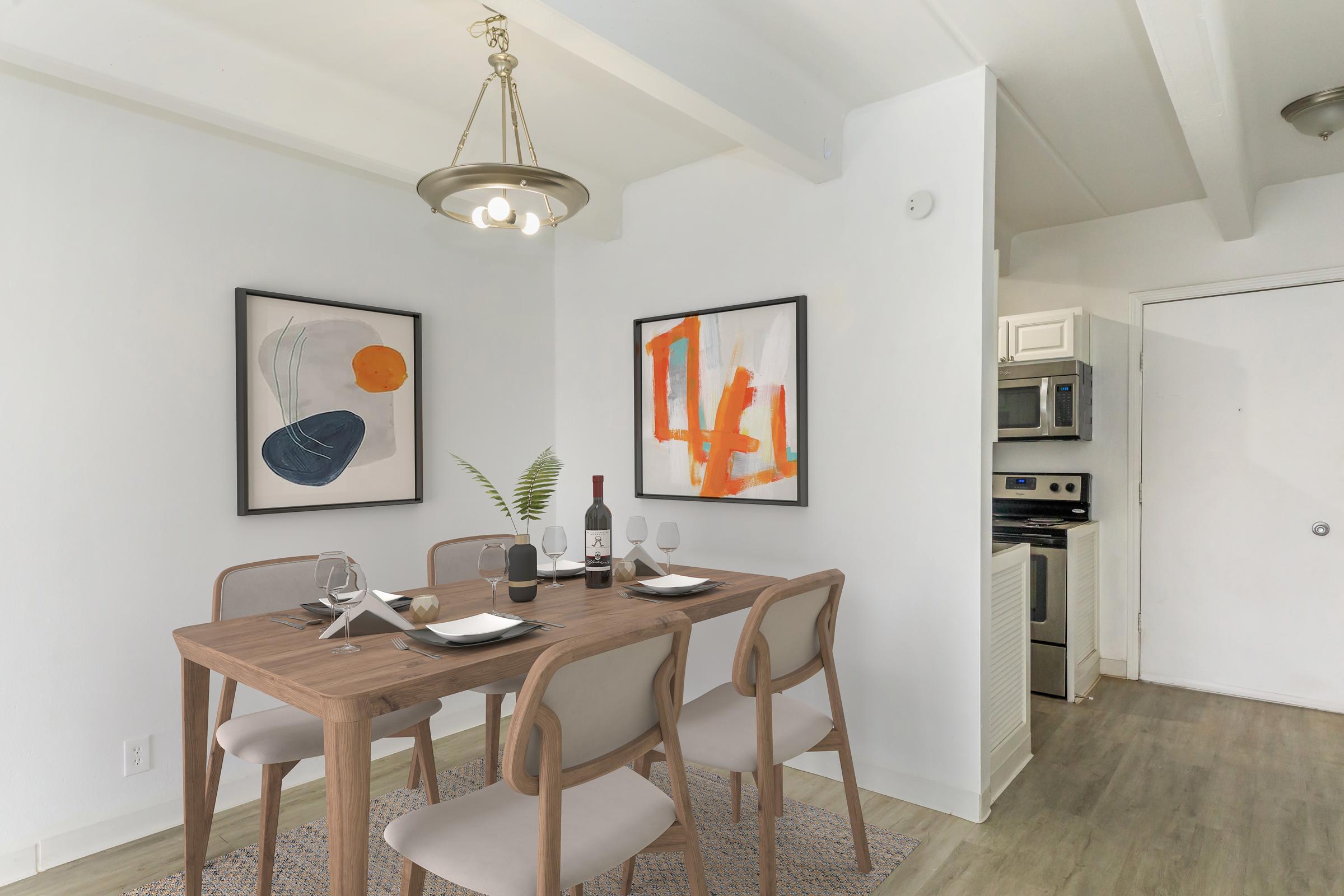 A modern dining area featuring a wooden table set for four with tableware and wine. On the walls, two contemporary art pieces in black, orange, and blue. A pendant light hangs above the table, and a small green plant adds a touch of nature. The kitchen is visible in the background with stainless steel appliances.