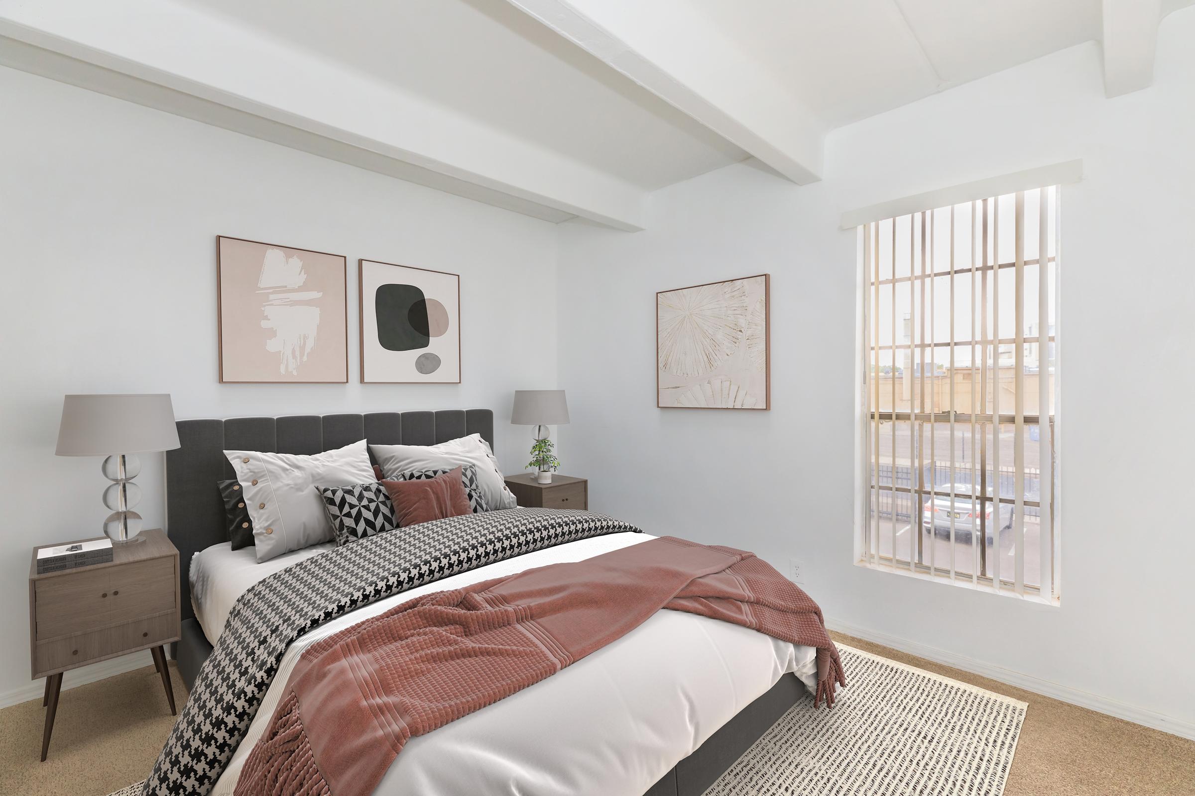 A stylish bedroom featuring a neatly made bed with a patterned throw and decorative pillows. The walls are adorned with abstract art, and there are modern bedside tables with lamps. A window with vertical blinds allows natural light to enter the room, creating a warm and inviting atmosphere.