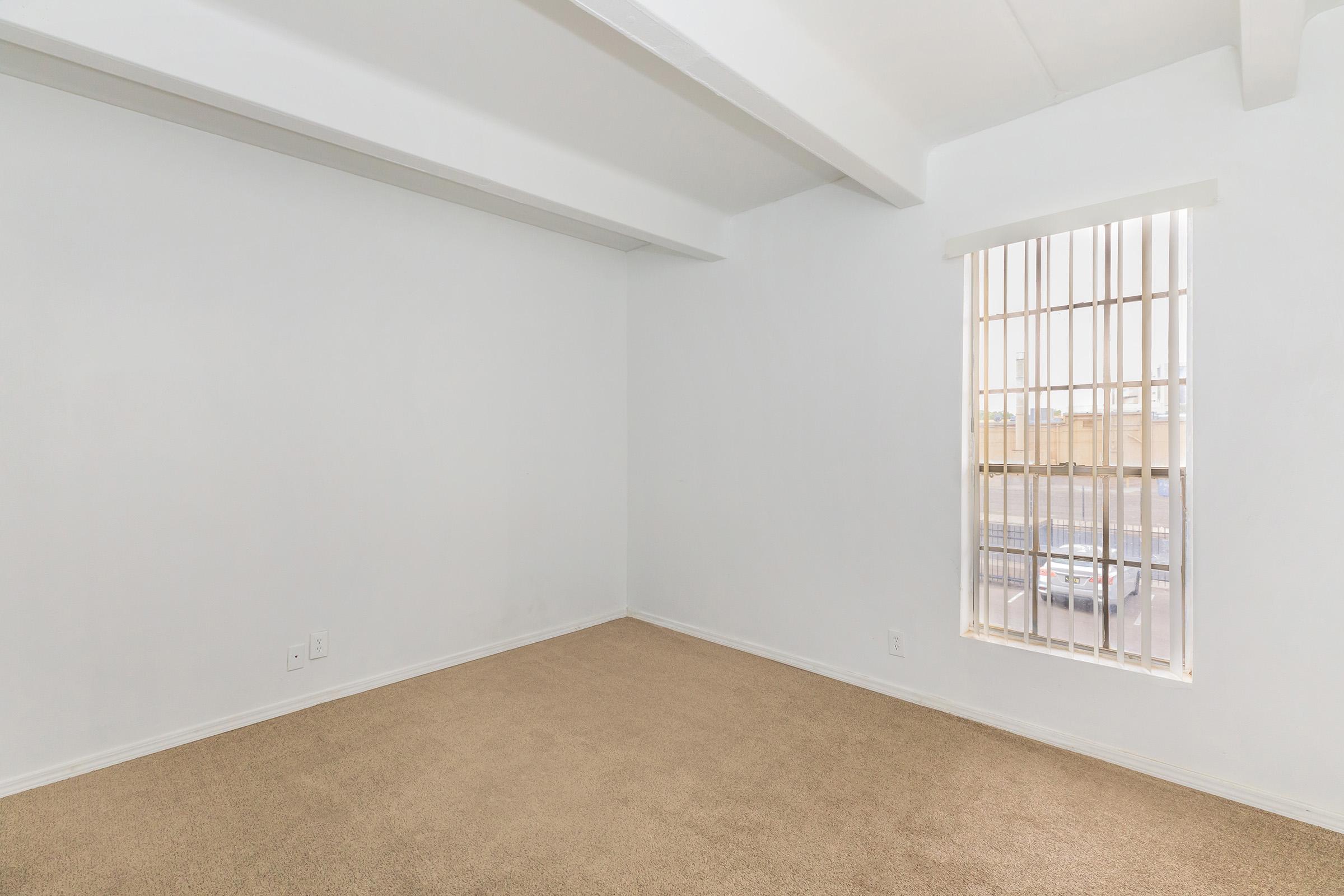Empty room with white walls and a tan carpet. It features a window with vertical blinds, allowing natural light to enter. The room has exposed ceiling beams, providing a spacious feel. No furniture or decorations present, creating a minimalist atmosphere.
