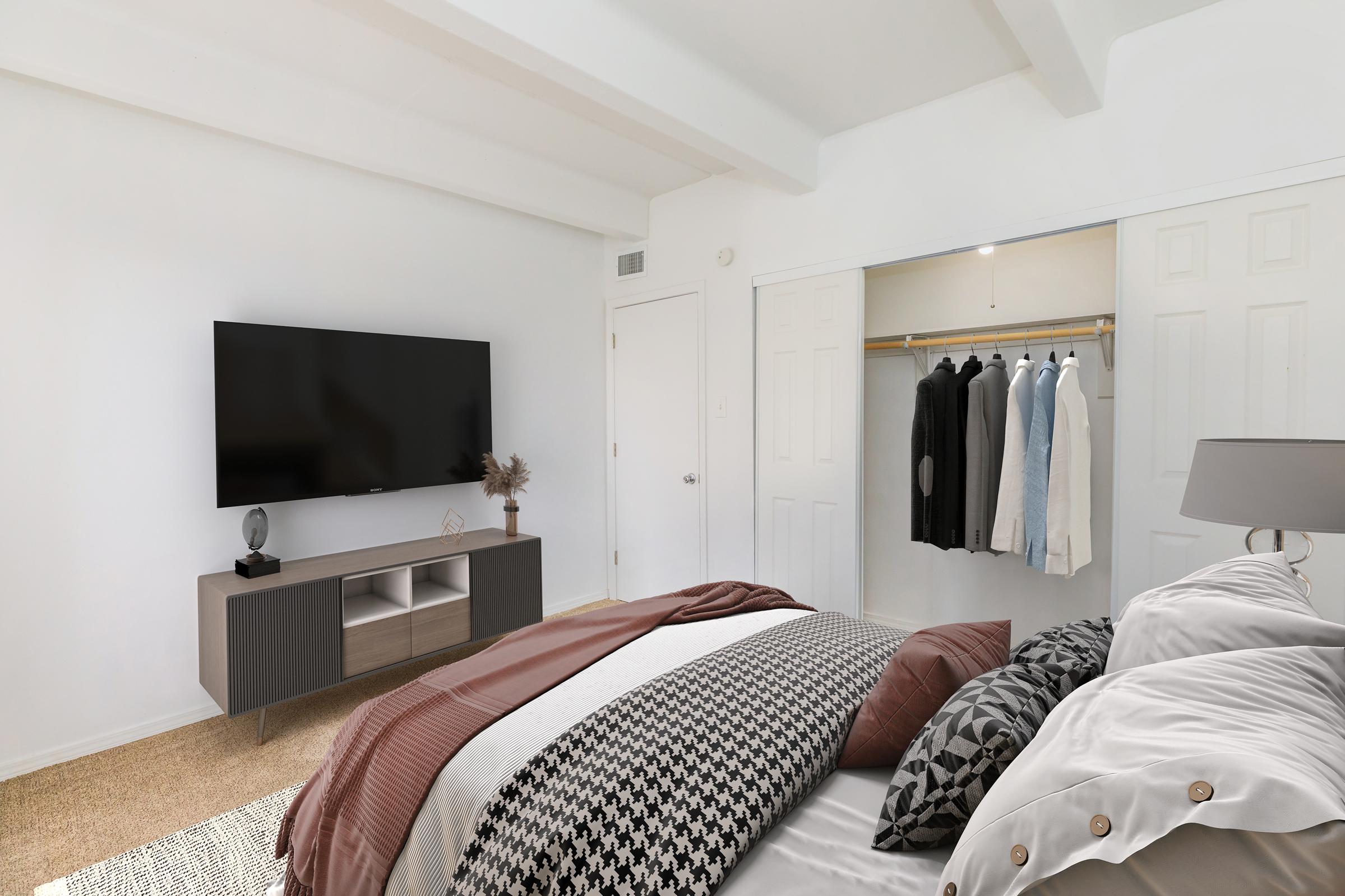 A modern bedroom featuring a neatly made bed with patterned blankets and pillows. A large flat-screen TV is mounted on the wall opposite the bed. There’s a wardrobe with hanging clothes, and a small decorative plant on a bedside table, all set against a bright, white wall.