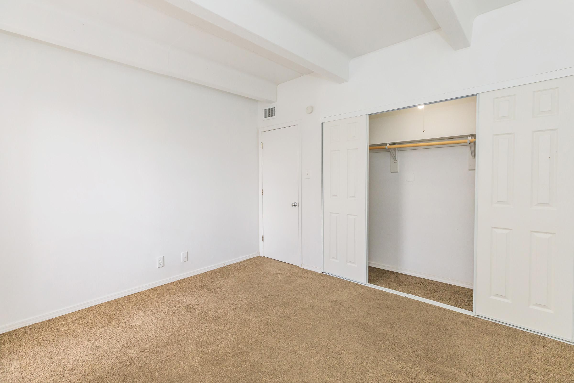 Empty room with light-colored walls and a tan carpet. There is a white door leading to a closet with double doors, which is open to reveal a wooden hanging rod. The room has a bright, spacious feel with no furniture or decorations visible.