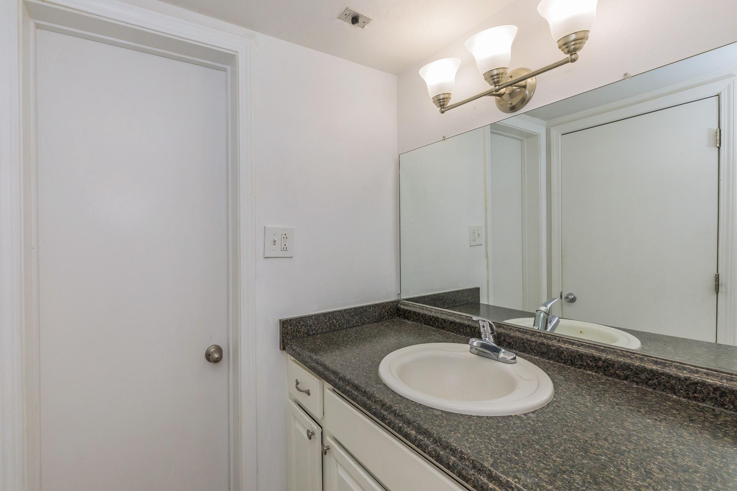 A clean and simple bathroom featuring a white countertop with a sink, a large mirror above it, and three light fixtures. There is a closed white door on the left and a second door on the right, both leading to separate spaces. The walls are painted a neutral color, enhancing the room's brightness.