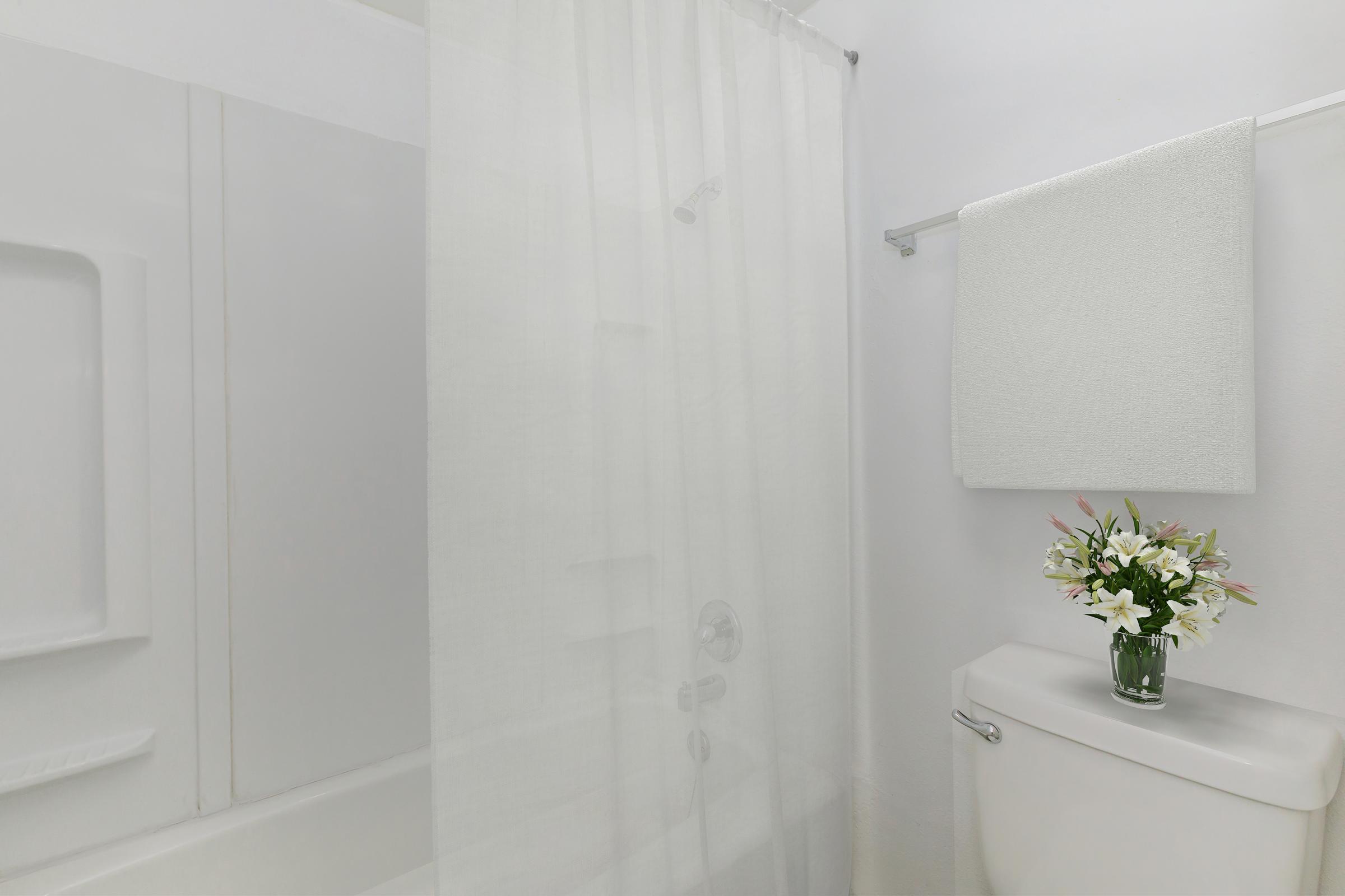 A bright, clean bathroom featuring a white shower curtain, a small towel hanging on a rod, and a vase of flowers on the toilet tank, creating a fresh and inviting atmosphere.