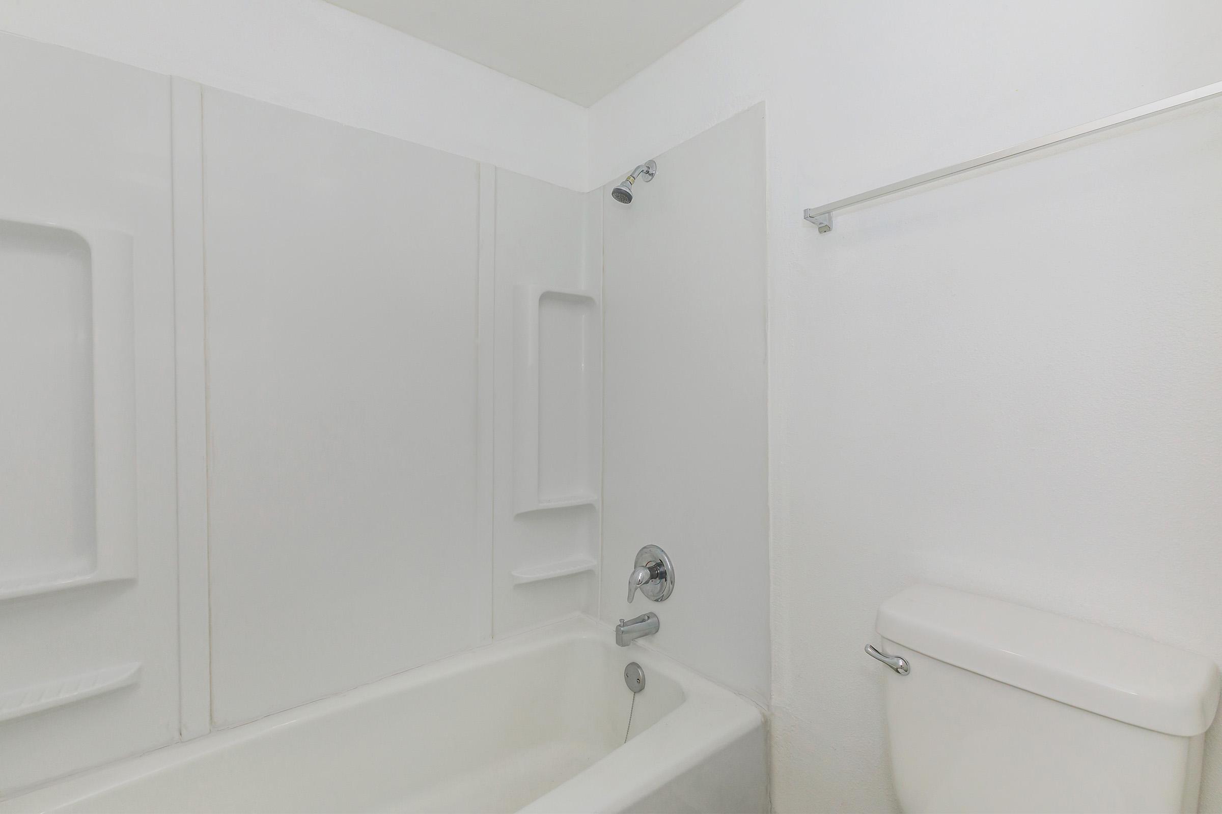 A clean and simple bathroom featuring a white bathtub, a showerhead mounted on the wall, and a toilet. The walls are plain white, and there is a towel bar installed. Overall, the space appears bright and minimalist.