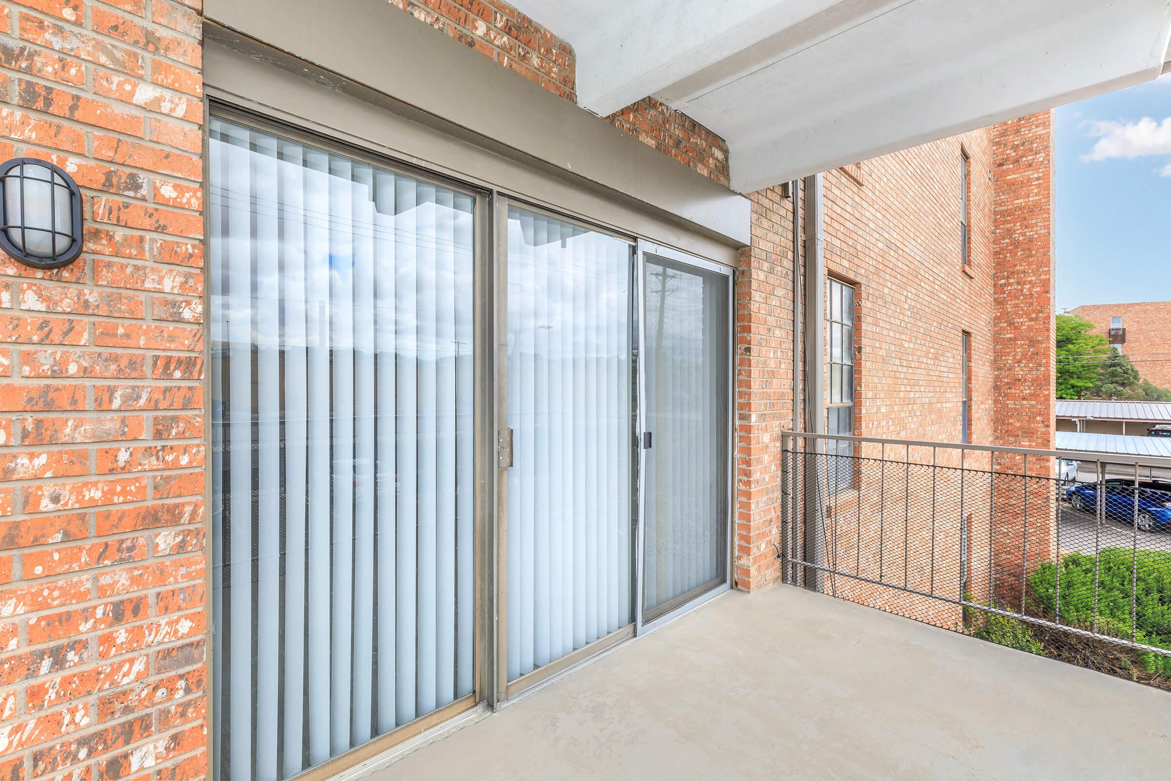 A balcony with a railing overlooks a brick building. Glass sliding doors with vertical blinds lead into a room. The floor is concrete, and the view includes a nearby building and a cloudy sky.