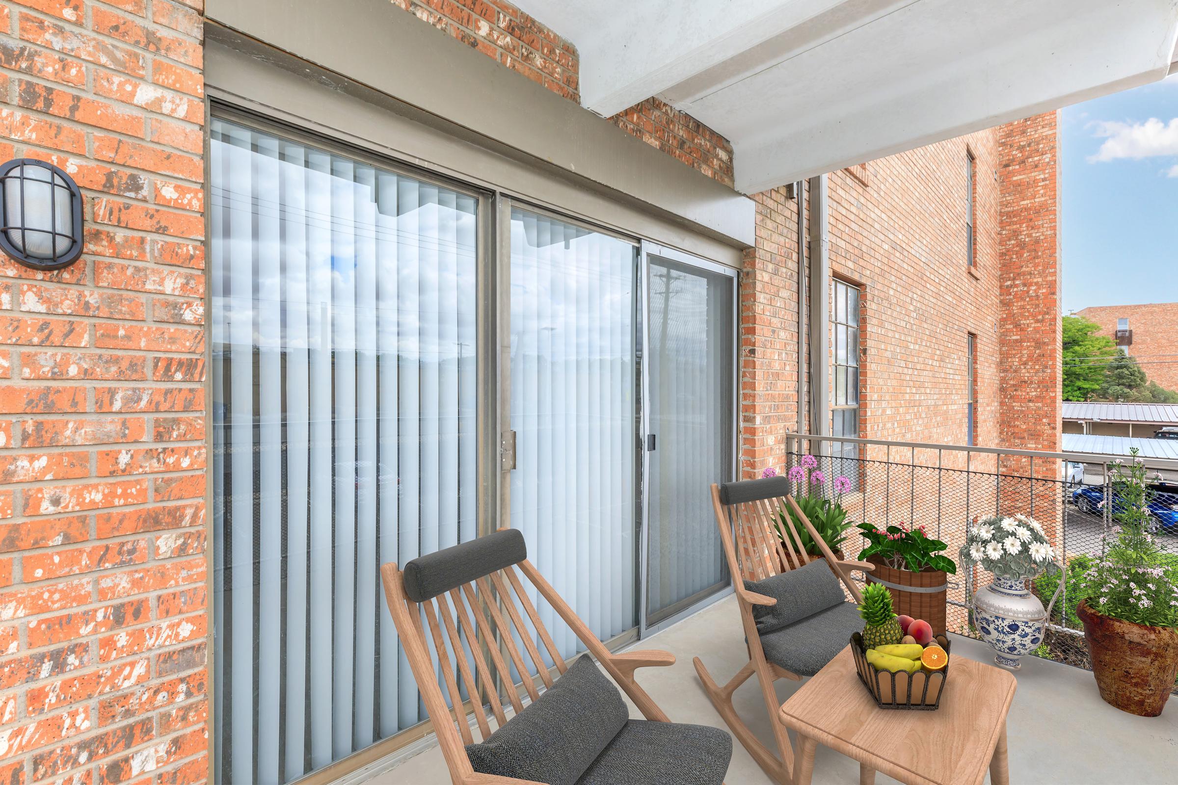 A cozy balcony featuring two wooden chairs and a small table with fruits. Potted plants and flowers are arranged nearby. The large sliding glass doors with vertical blinds reflect the surroundings and provide a view of the outside. The brick wall adds a warm, rustic touch to the space.
