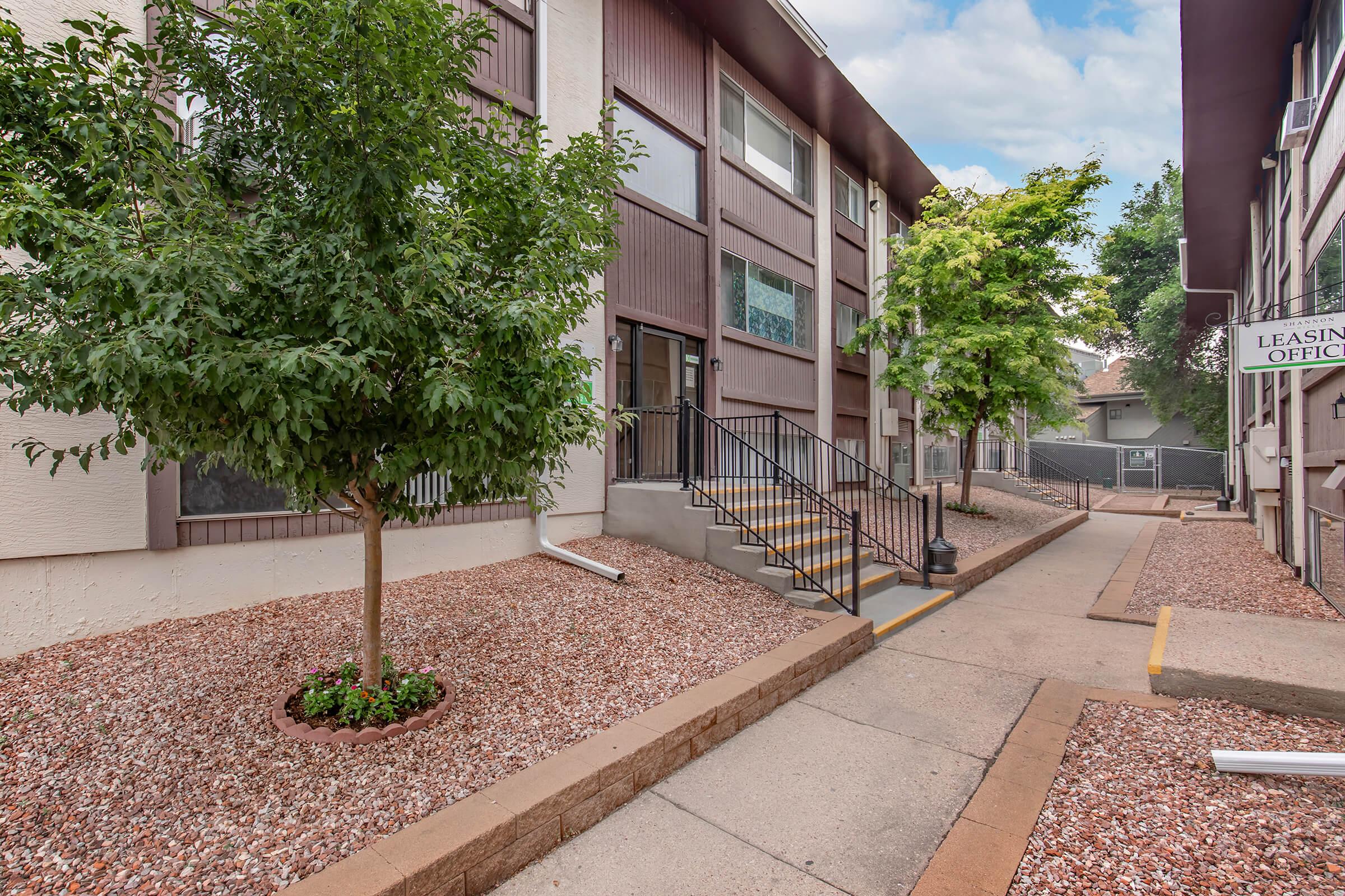 A pathway between two apartment buildings, featuring landscaped areas with small trees and gravel. Stairs lead up to an entrance, and a leasing office sign is visible in the background. The scene is bright and well-maintained, with a blue sky overhead and green foliage.