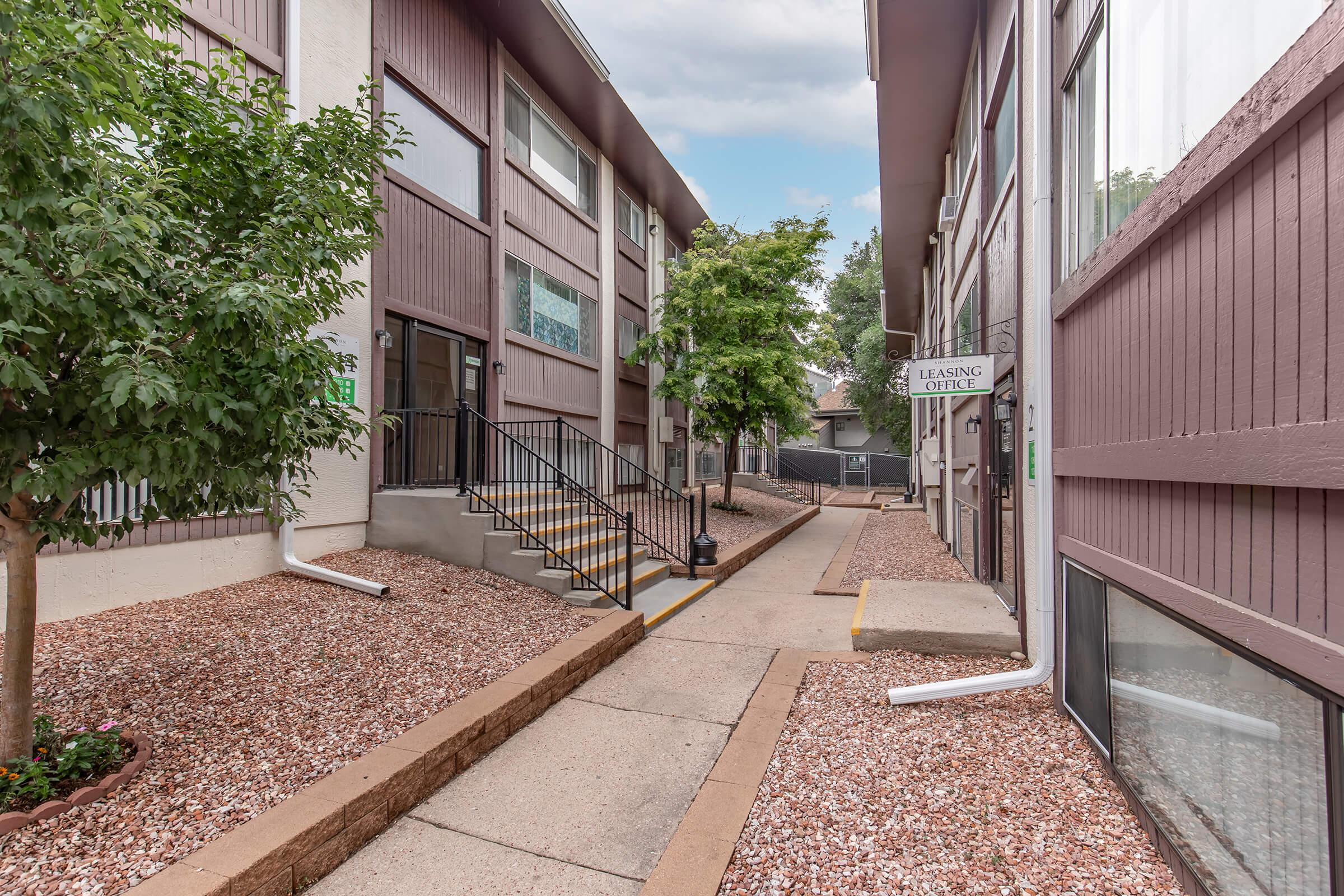 A view of a residential complex pathway lined with gravel and small trees. The path leads to a building with a "Leasing Office" sign. Stairs are visible, and the surrounding buildings have large windows. The scene is well-lit, suggesting a clear day.