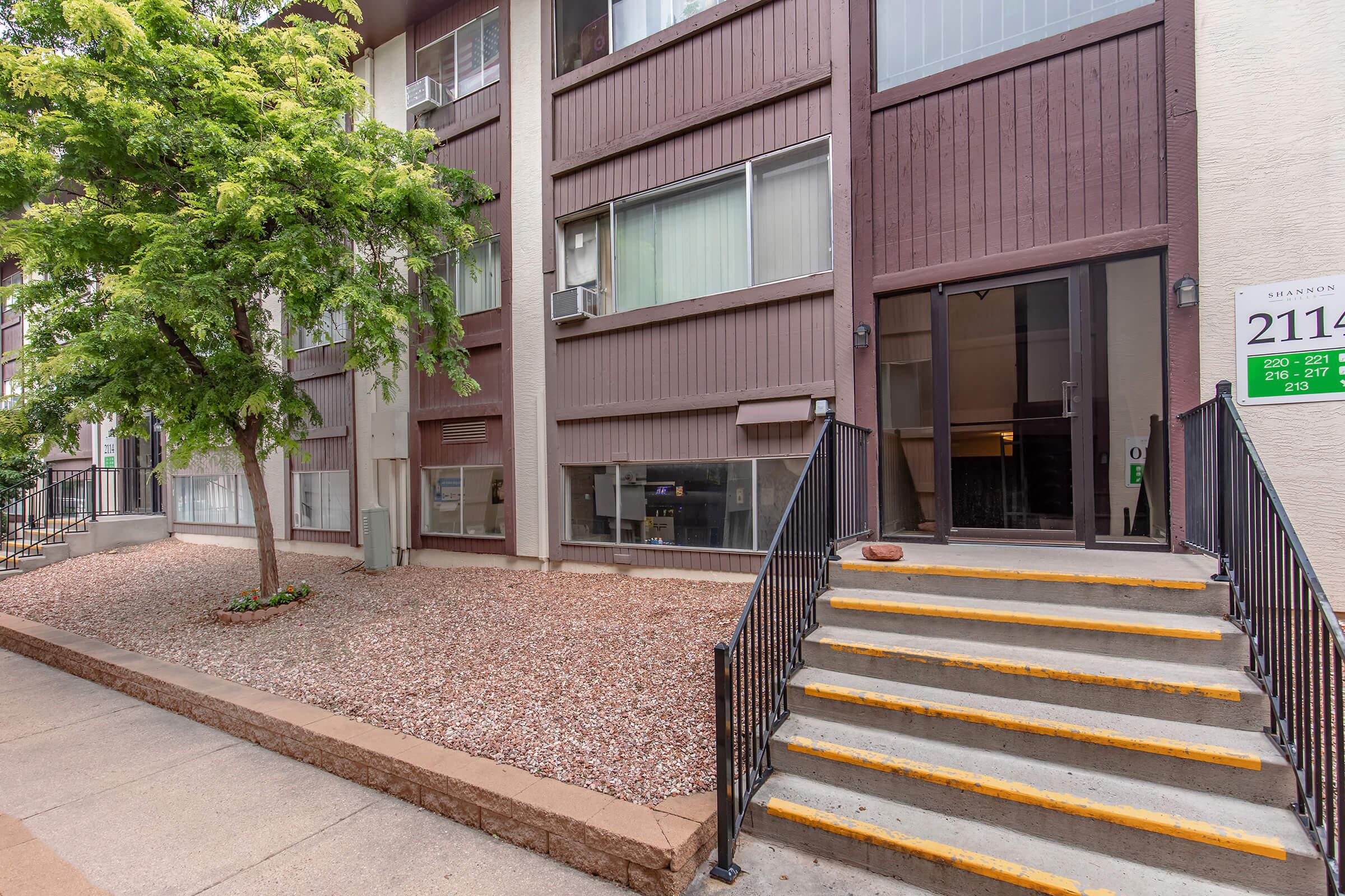 A three-story residential building with a brown exterior. The entrance features a set of yellow-edged stairs leading up to the door. To the left, there is a small landscaped area with a tree and gravel. Windows are visible on the ground and upper floors, and there is a sign near the entrance with property information.