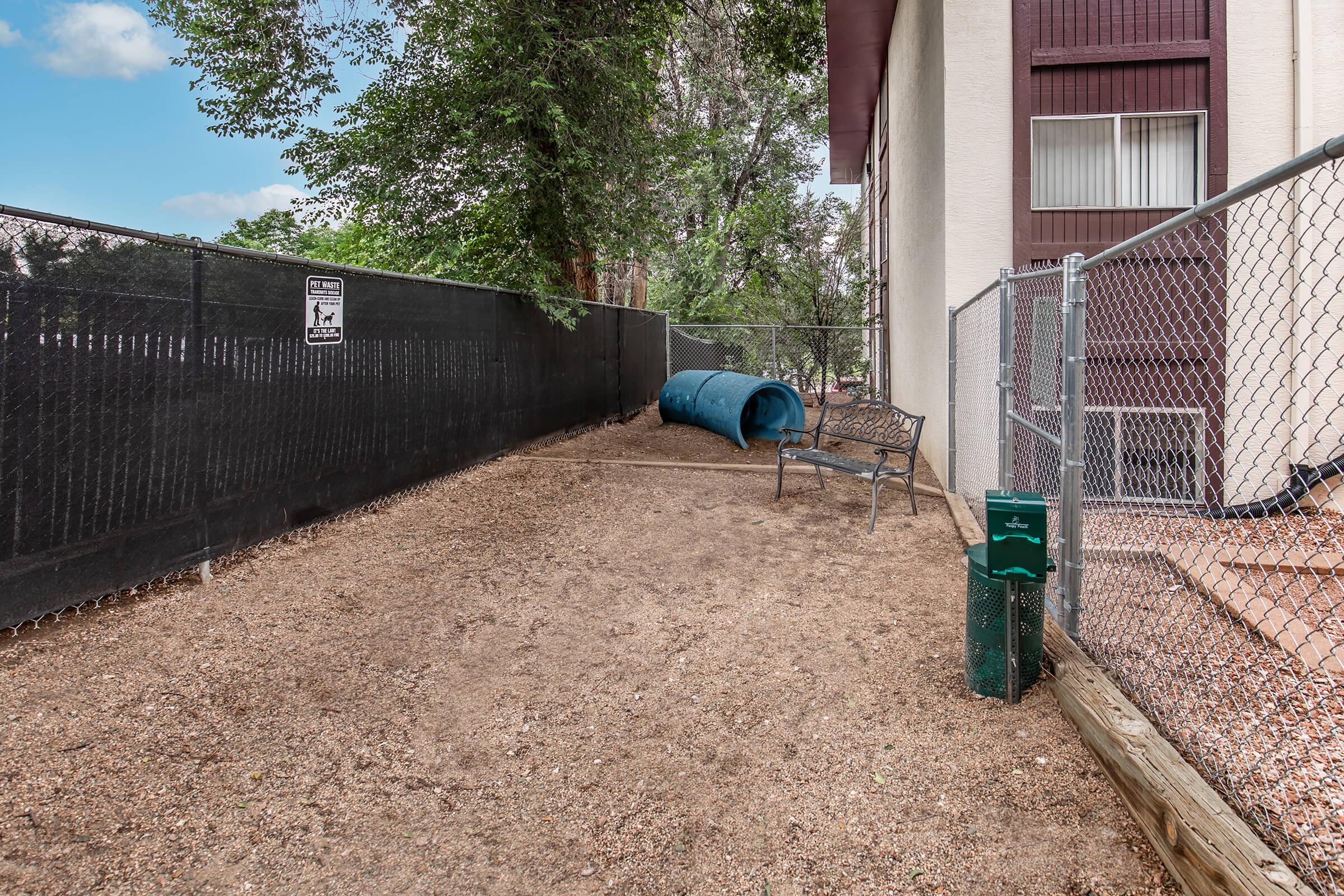 A fenced outdoor area with gravel flooring, featuring a blue tunnel, a wooden bench, and a waste disposal station. There are trees in the background and a building structure nearby. The environment is designed for pets, likely for use as a dog park or play area.