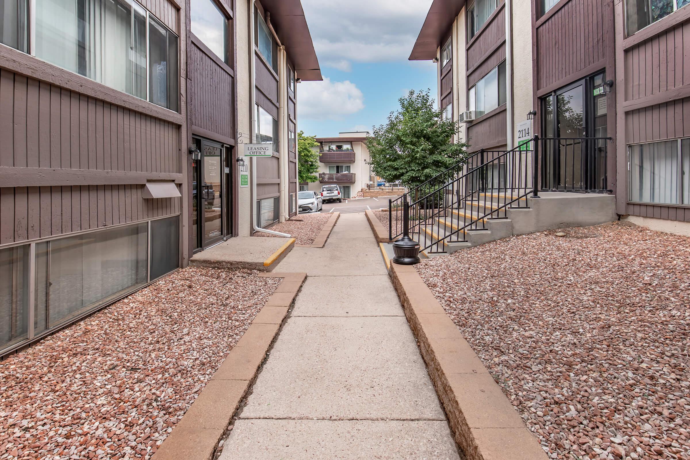 A narrow, paved walkway between two multi-story apartment buildings, lined with gravel. On one side, there are staircases leading to individual units, while on the other, trees and parking can be seen in the background under a partly cloudy sky.