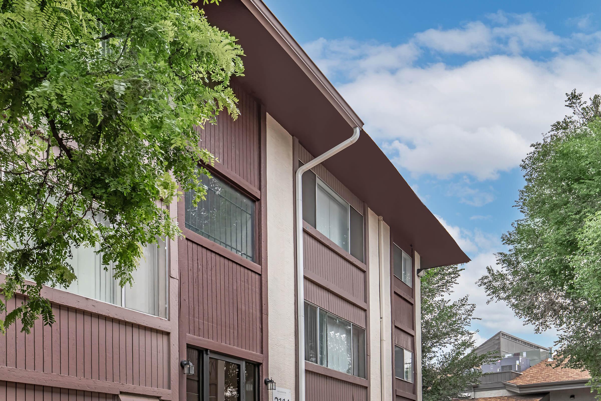 A close-up view of a modern apartment building featuring brown wooden paneling and large windows. The image shows a portion of the building against a bright blue sky with scattered clouds, framed by green foliage from nearby trees.