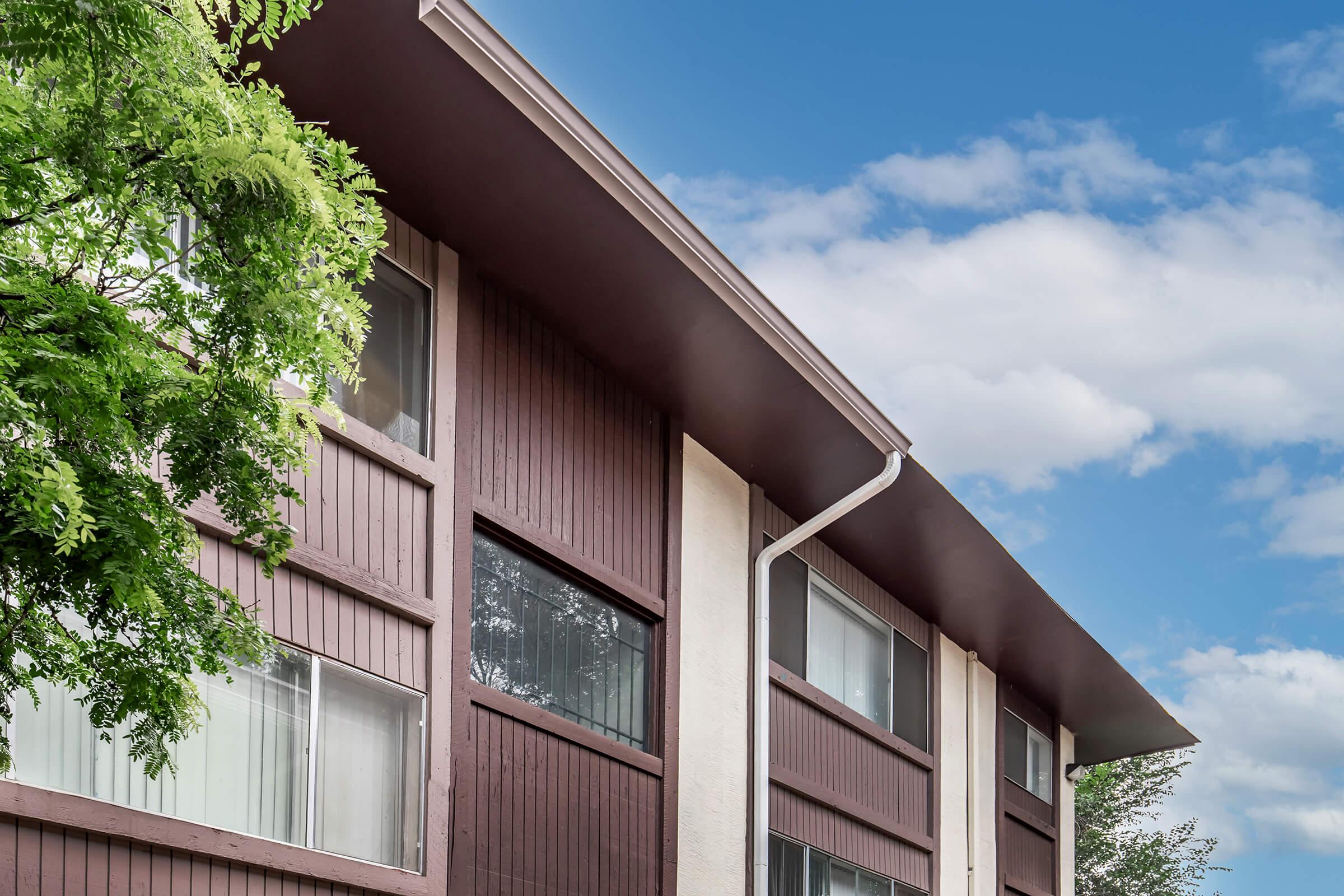 A close-up view of a modern building facade with wooden paneling and large windows, set against a partly cloudy blue sky. Lush green foliage is visible in the foreground, adding a natural element to the scene. The angle captures the roofline and architectural details of the structure.