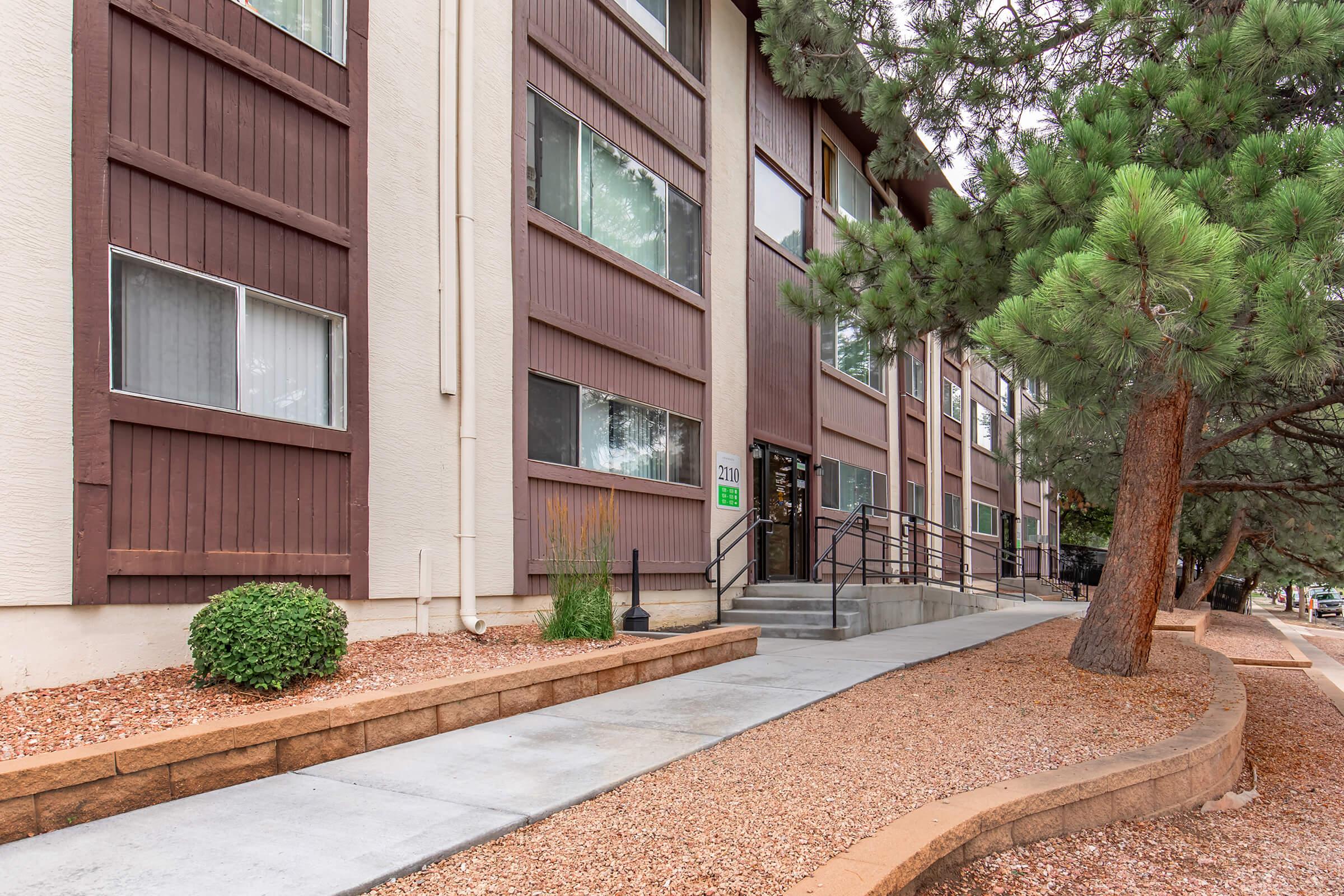 Exterior view of a modern building with a light-colored facade and large windows, featuring a landscaped walkway with gravel and shrubs leading up to the entrance. There are steps and a railing, with a few trees nearby, creating a welcoming atmosphere.