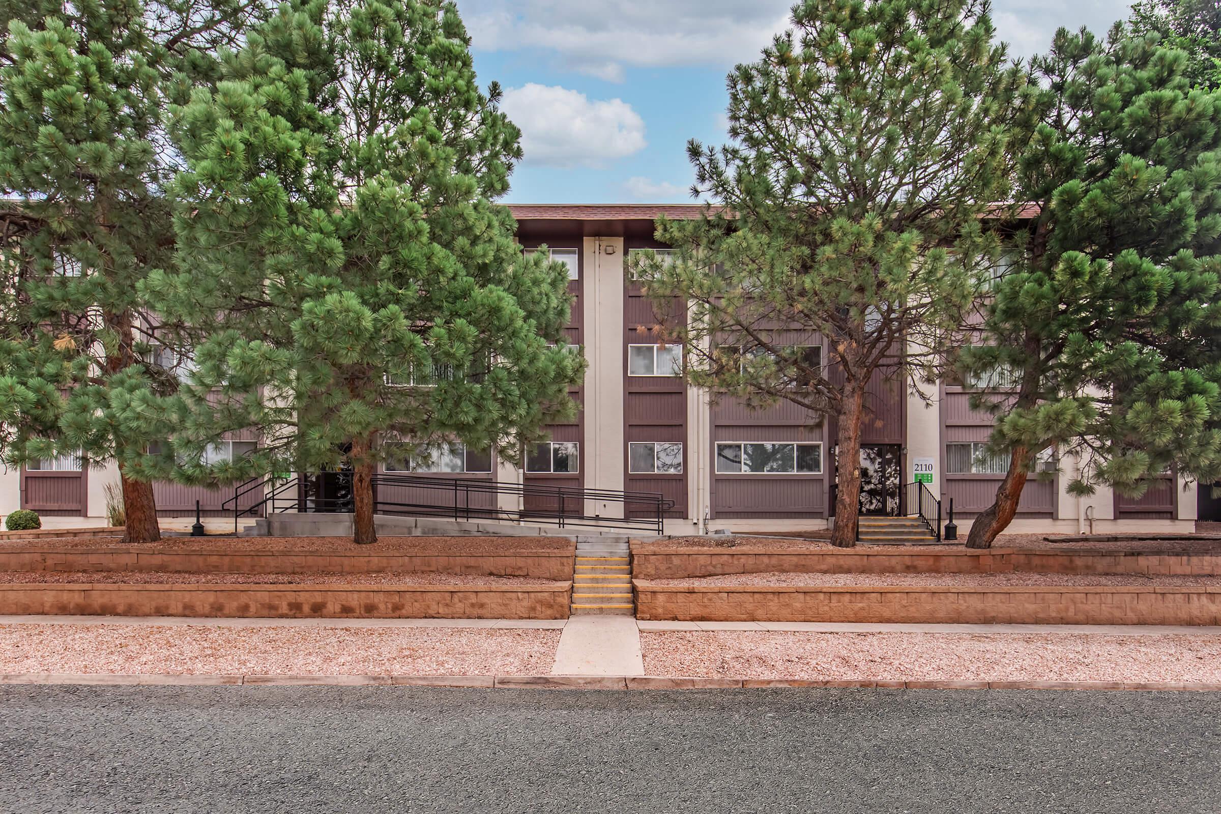 A residential building surrounded by tall pine trees, with a front walkway leading up to the entrance. The structure features large windows and a mix of brown and beige colors. A gravel area is in front of the building, flanked by landscaped greenery under a partly cloudy sky.