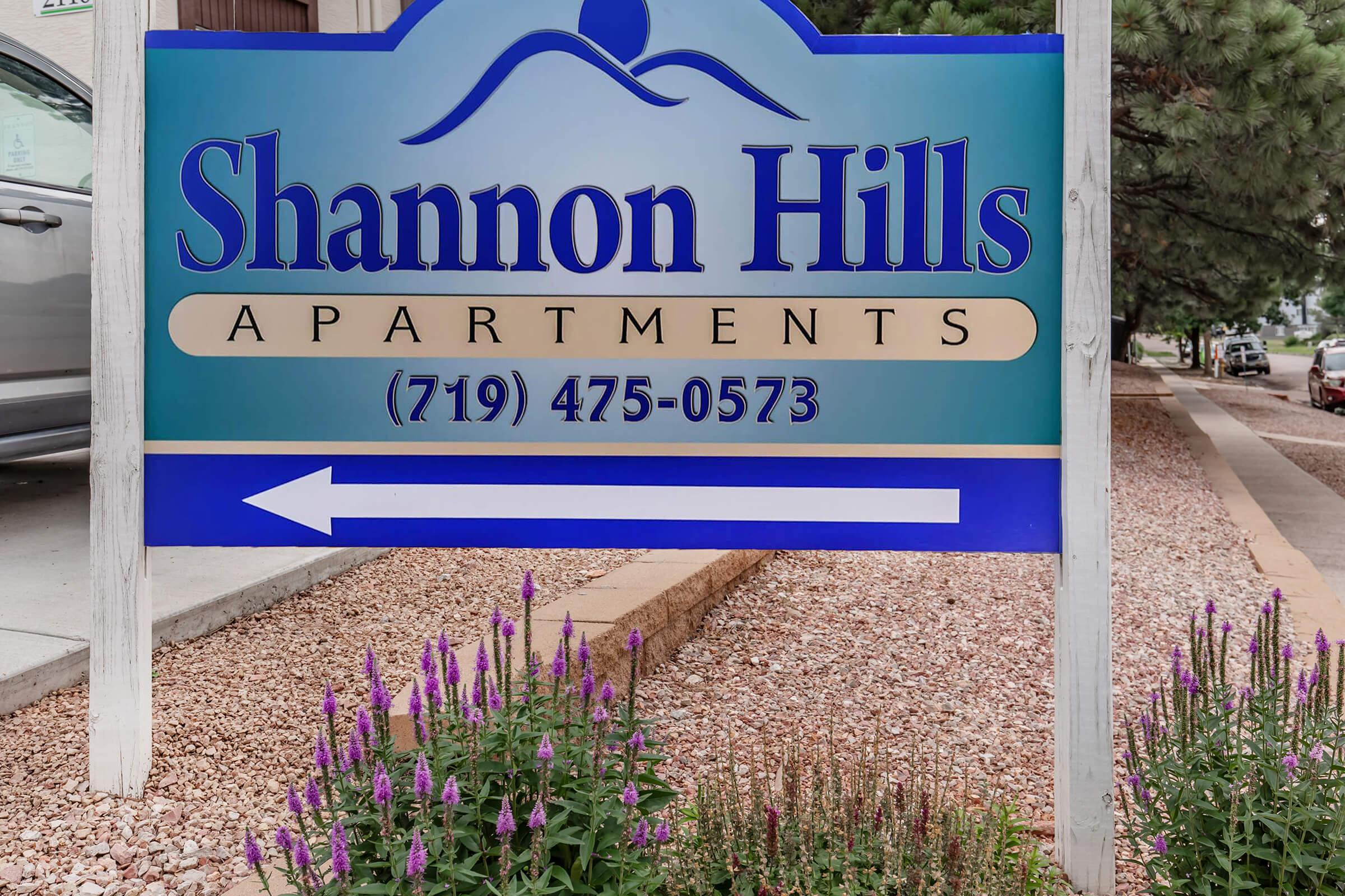 Sign for Shannon Hills Apartments with blue and white lettering, featuring a decorative wave design. Below the name is the phone number (719) 475-0573. A directional arrow points left. Surrounding the sign are purple flowers and stones, indicating a landscaped area.