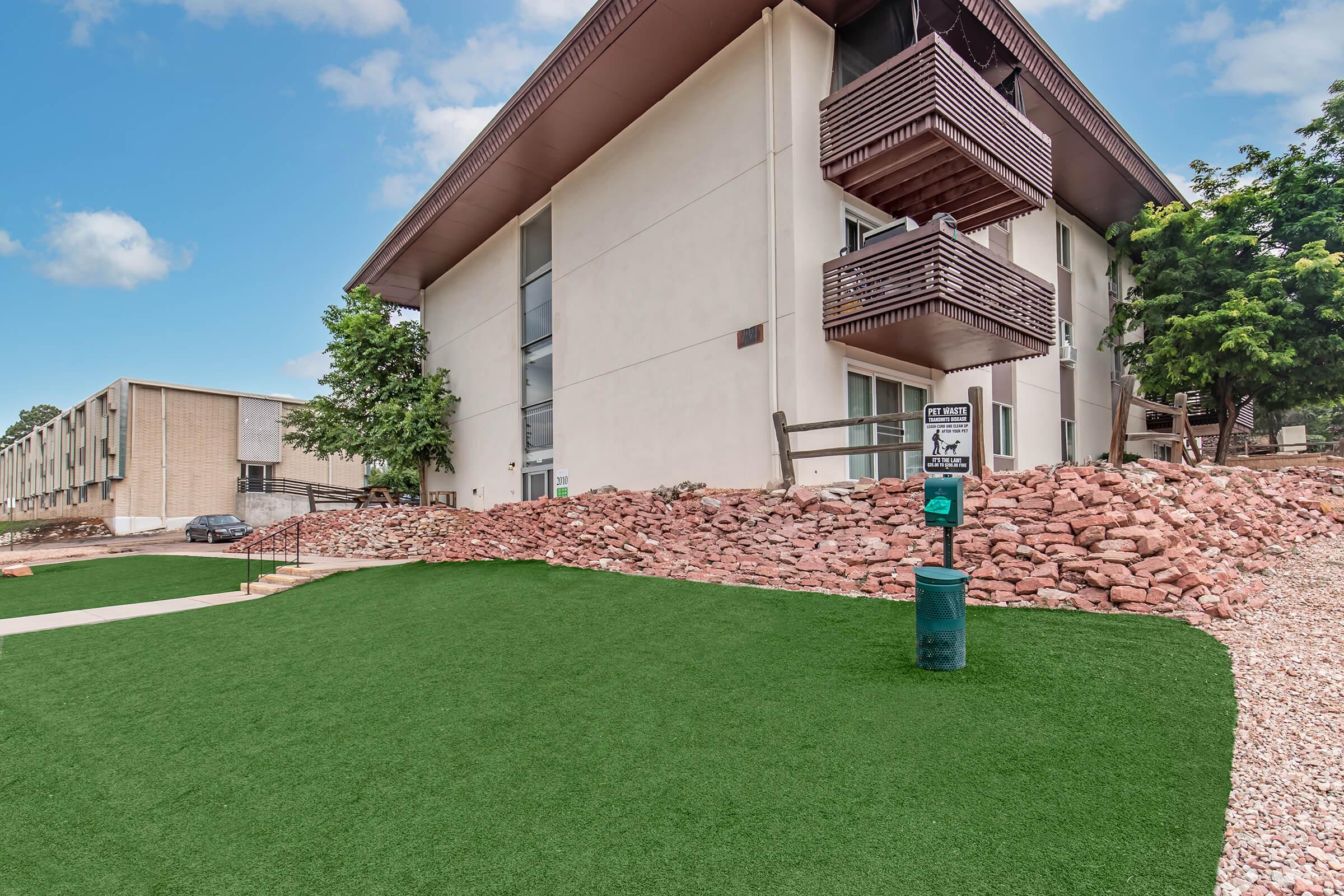 Apartment building with a modern design featuring balconies and large windows. The foreground has a green, manicured lawn, with a stone pathway leading to the entrance. Nearby, a park bench and a visible sign for dog waste disposal can be seen. The background includes a second building, set against a blue sky with clouds.