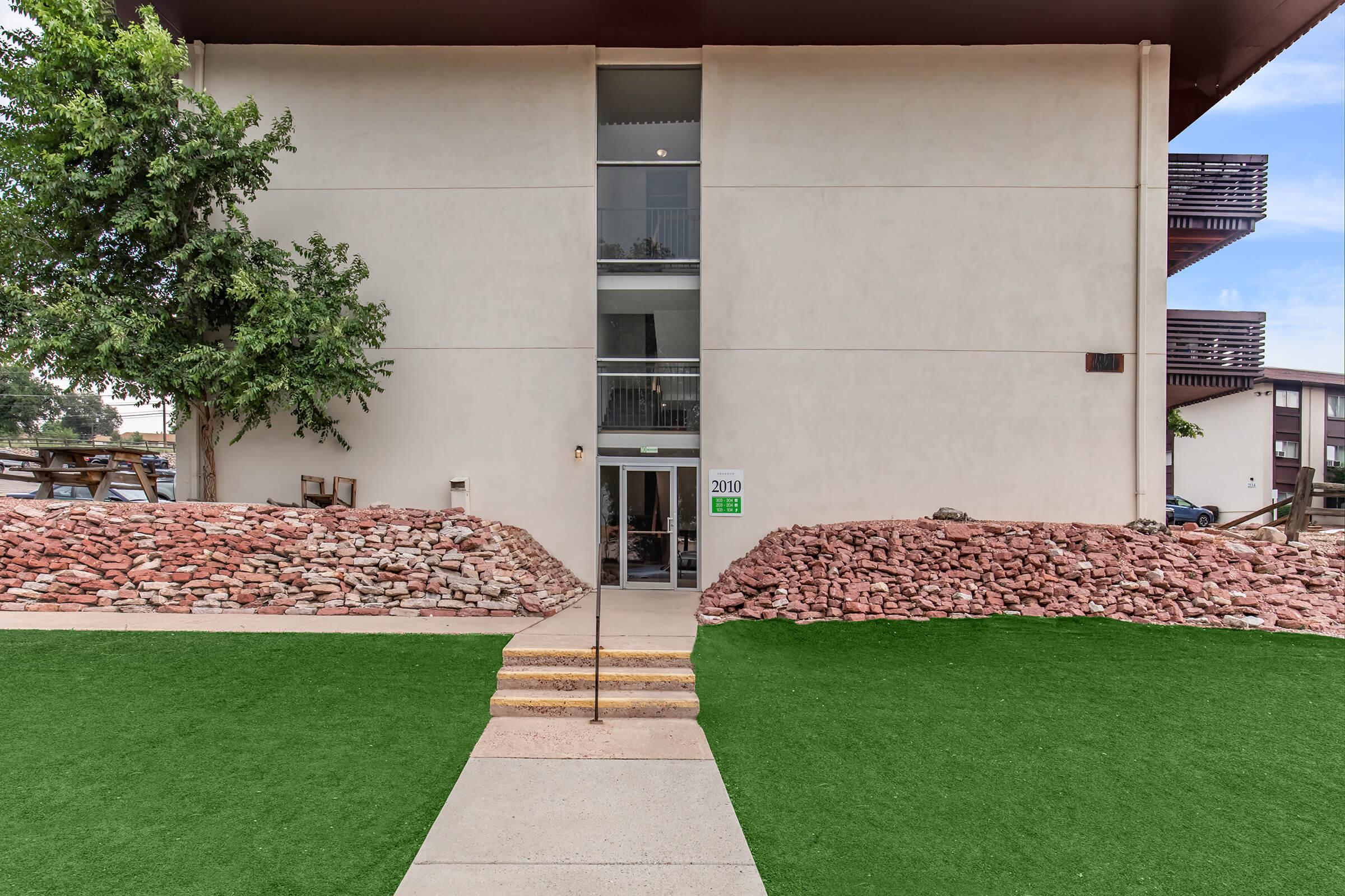 Entrance to a modern building with a large glass entrance door. Flanked by a landscaped area featuring rock formations and green grass. The building has a minimalist design with a second story visible above. A small set of stairs leads up to the entrance.