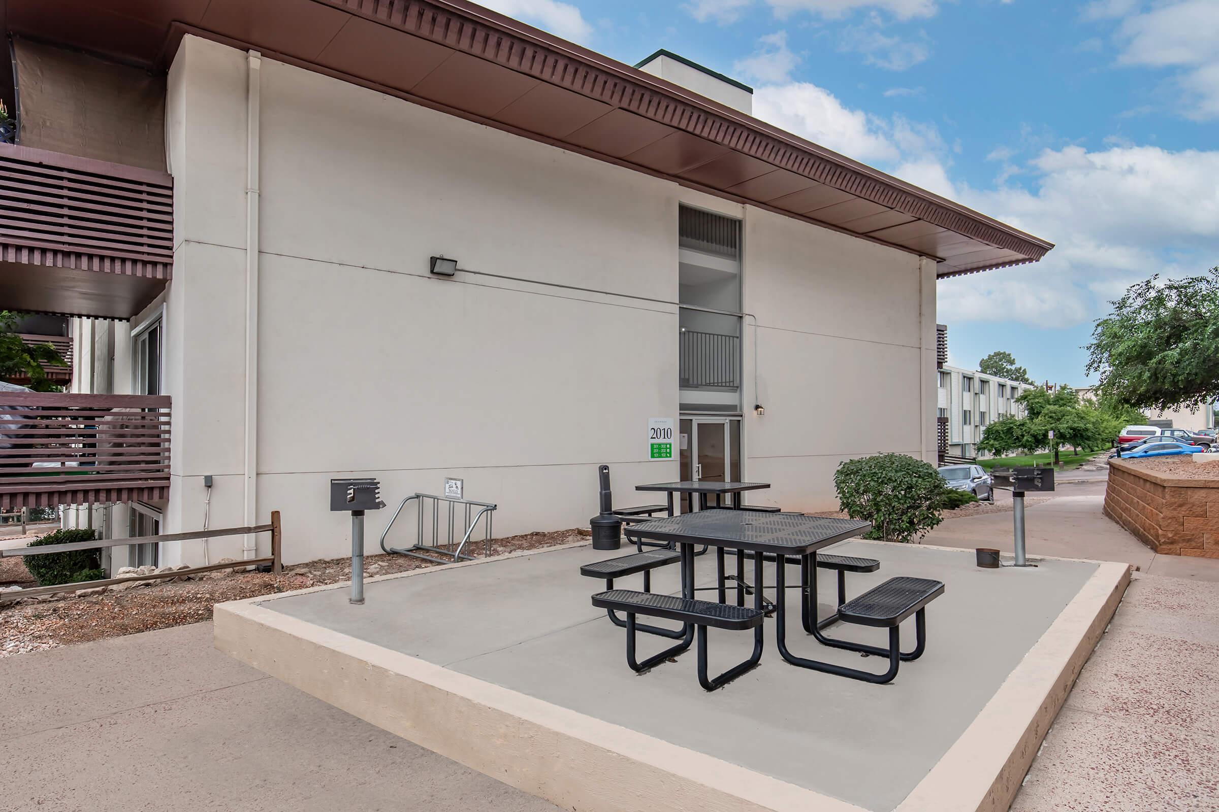 Outdoor area featuring a concrete patio with a picnic table and benches, surrounded by greenery. A large building with a modern design stands in the background, and there are additional amenities like grills nearby. The scene is well-lit with partly cloudy skies above.