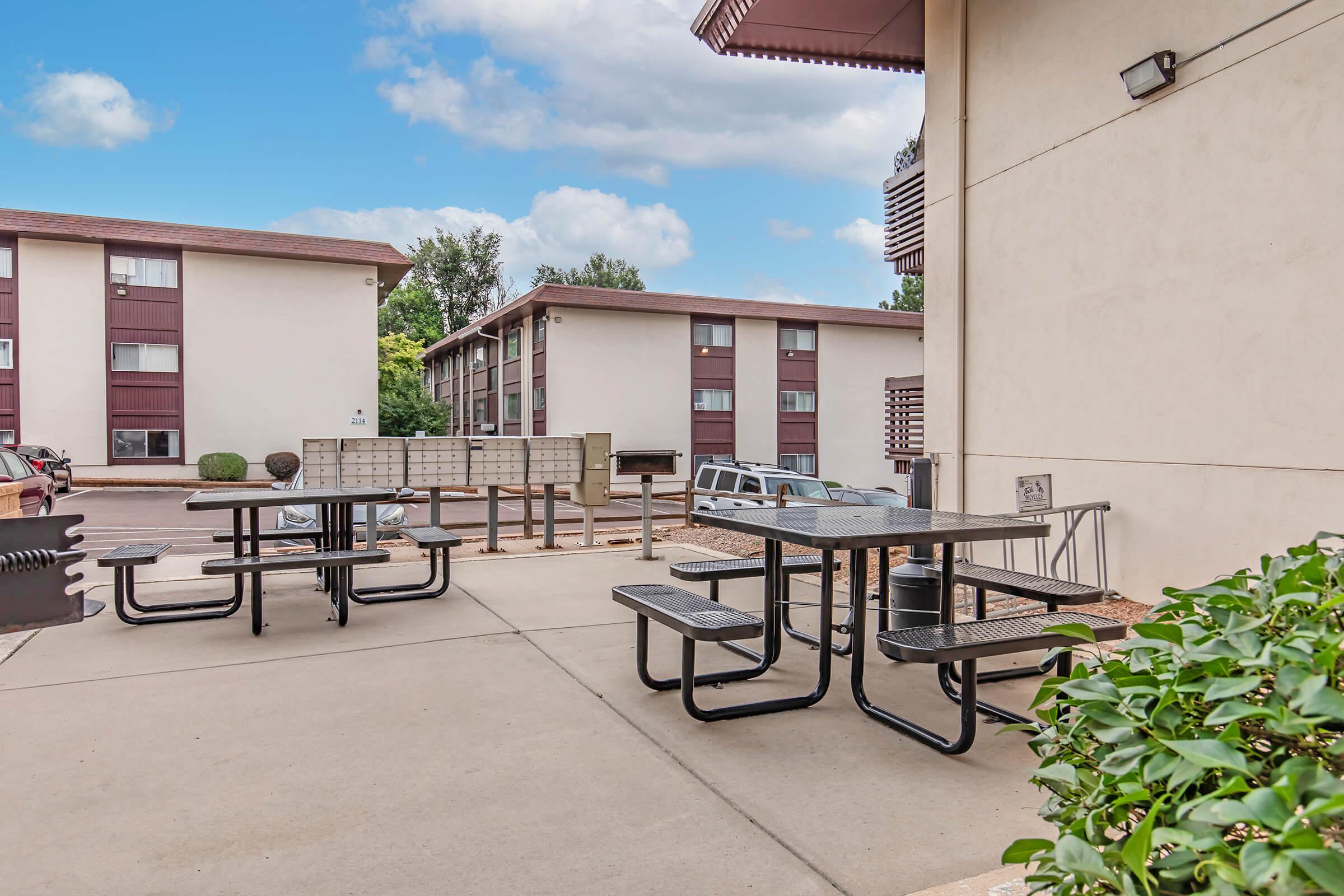 An outdoor area featuring a black picnic table with benches, surrounded by a concrete pathway. In the background, there are two multi-story apartment buildings and a row of mailboxes. The sky is partly cloudy, and there is some greenery visible near the edge of the image.