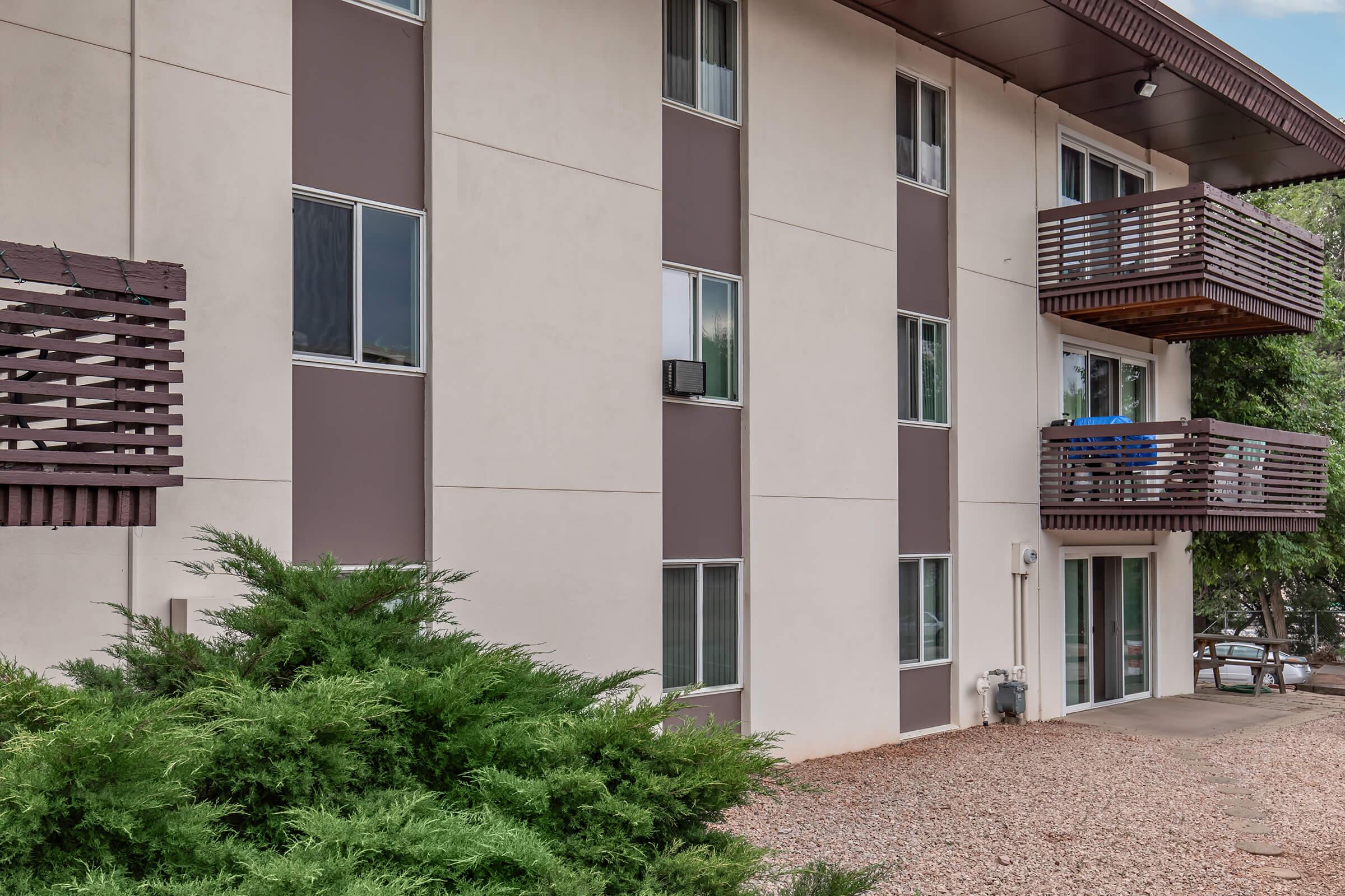 Exterior view of a multi-story apartment building featuring a modern design with a mix of light and dark wall panels. The building has several windows and small balconies with railing. In the foreground, there's a lush green shrubbery and gravel landscaping.