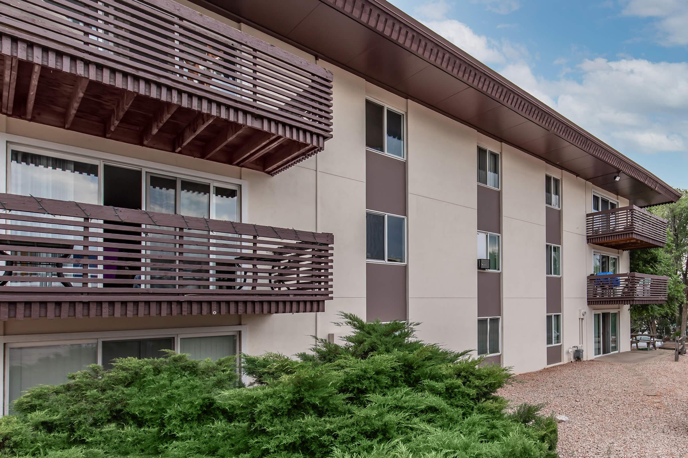 Three-story apartment building with a beige exterior and brown wooden balconies. The ground level features a landscaped area with bushes. Windows are visible on each floor, some partially obscured by curtains. The sky is partly cloudy, and gravel is present in the foreground.