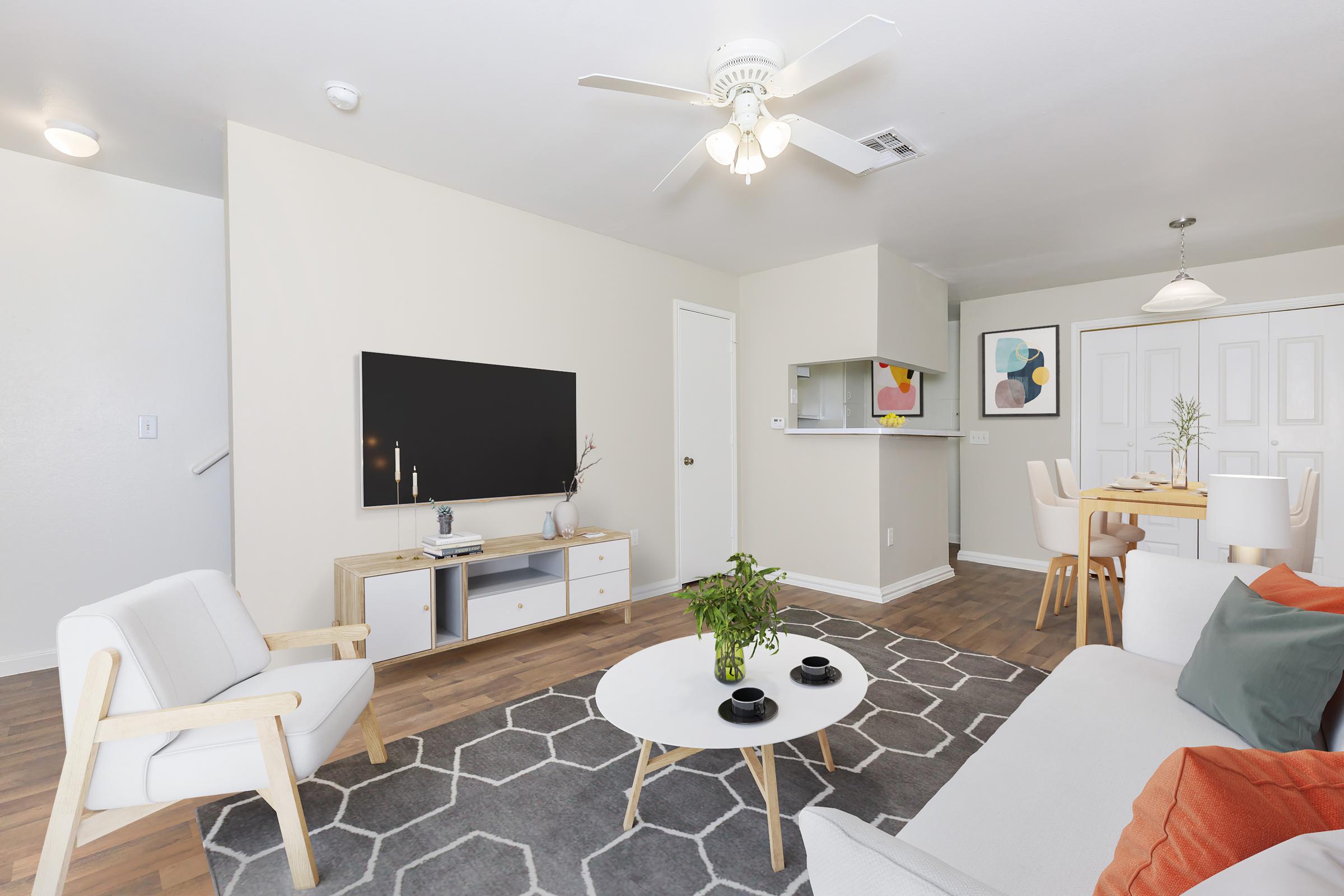 A modern living room featuring a white sofa with colorful cushions, a wooden armchair, and a large flat-screen TV on a white media console. A geometric gray area rug covers the floor, and a dining area with a light wood table and chairs is visible in the background, complemented by a ceiling fan and soft lighting.