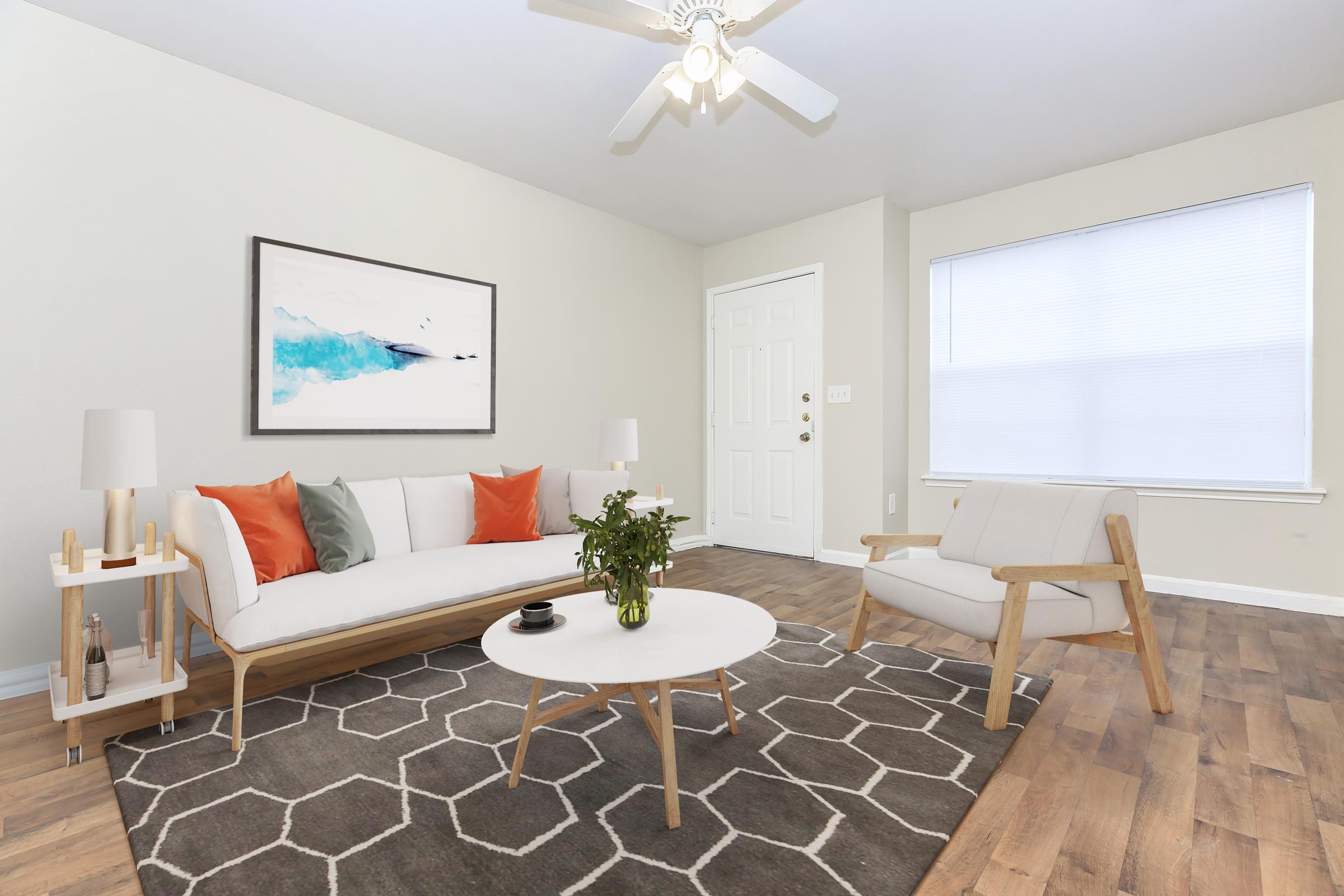 A bright and inviting living room featuring a white sofa with orange and gray cushions, a round coffee table on a geometric rug, and a comfortable chair. Natural light comes through a large window, and a piece of abstract artwork hangs on the wall, enhancing the modern decor.