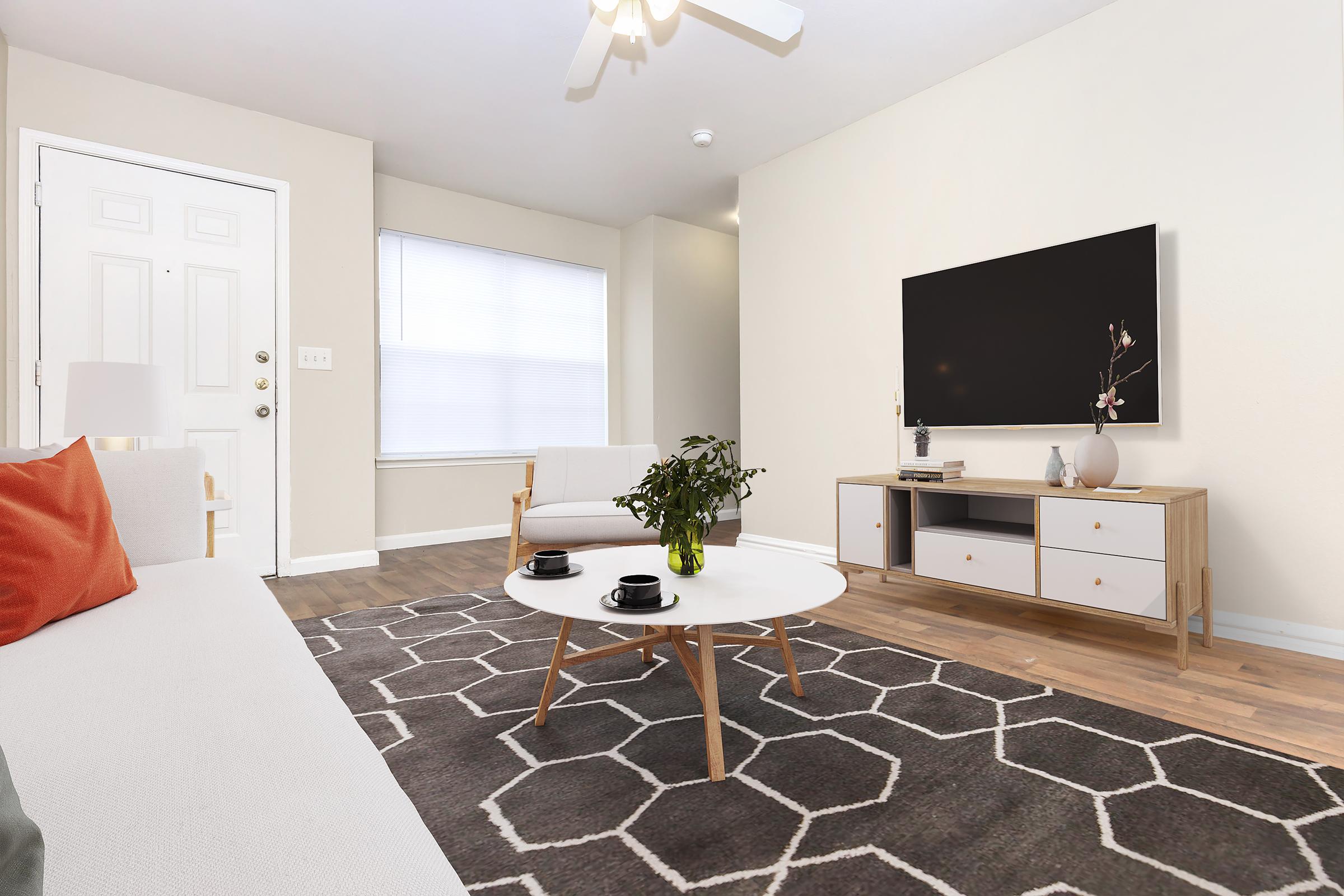 A modern living room featuring light-colored walls, a white sofa with an orange pillow, and a stylish coffee table with two cups. A flat-screen TV is mounted on the wall opposite the sofa, and there's a small plant on the coffee table. The space has wooden flooring and a geometric area rug. Natural light comes through a window.